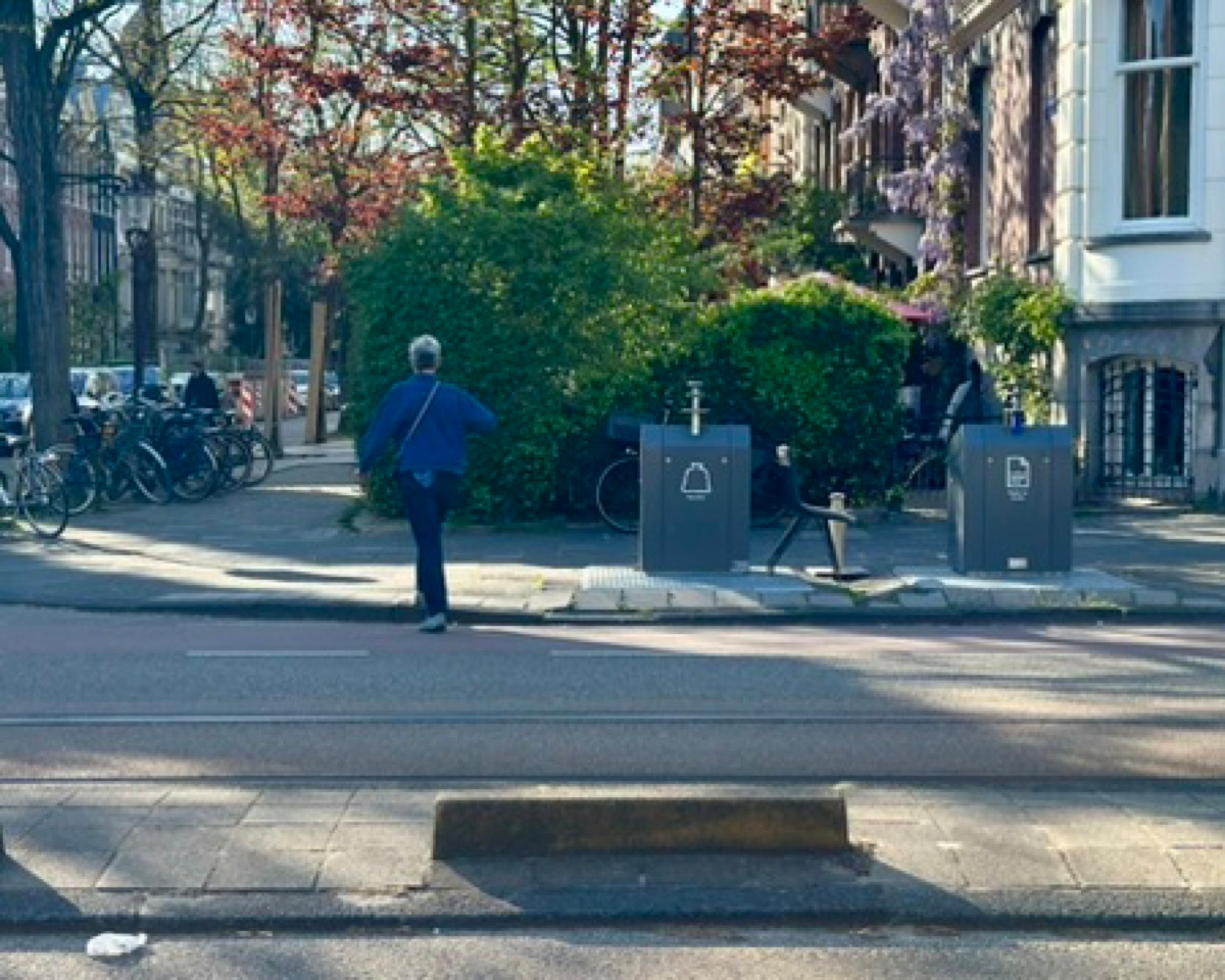 Amsterdam street scene: a person in a blue jacket walks near two underground waste containers. Bikes parked along a railing, wisteria blooming on a building to the right, a stone block in the foreground.