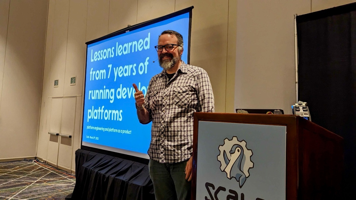 Coté at the SCALE conference podium, thumbs up, in front of a slide titled 'Lessons learned from 7 years of running developer platforms.'