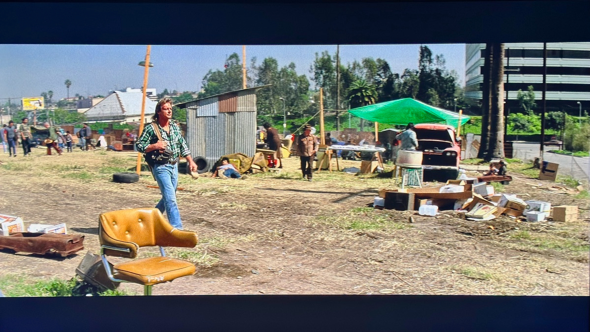 A yellow vinyl office swivel chair abandoned in a vacant lot from the movie They Live, with Roddy Piper walking past in a plaid shirt