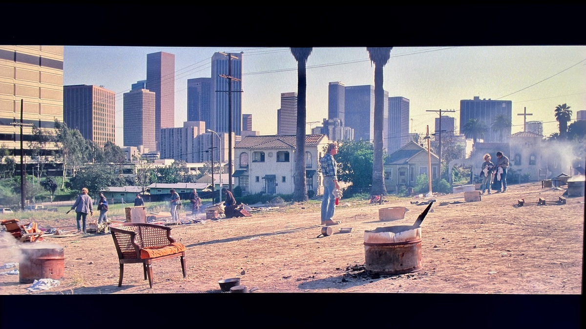 A wicker chair with a cushion sits in a dusty vacant lot from the movie They Live, with the downtown LA skyline in the background