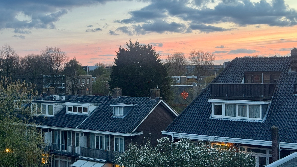 Sunset over Duivendrecht rooftops with pink and orange sky, dark tiled roofs, dormers, and a neon flame sign on the fire station in the distance