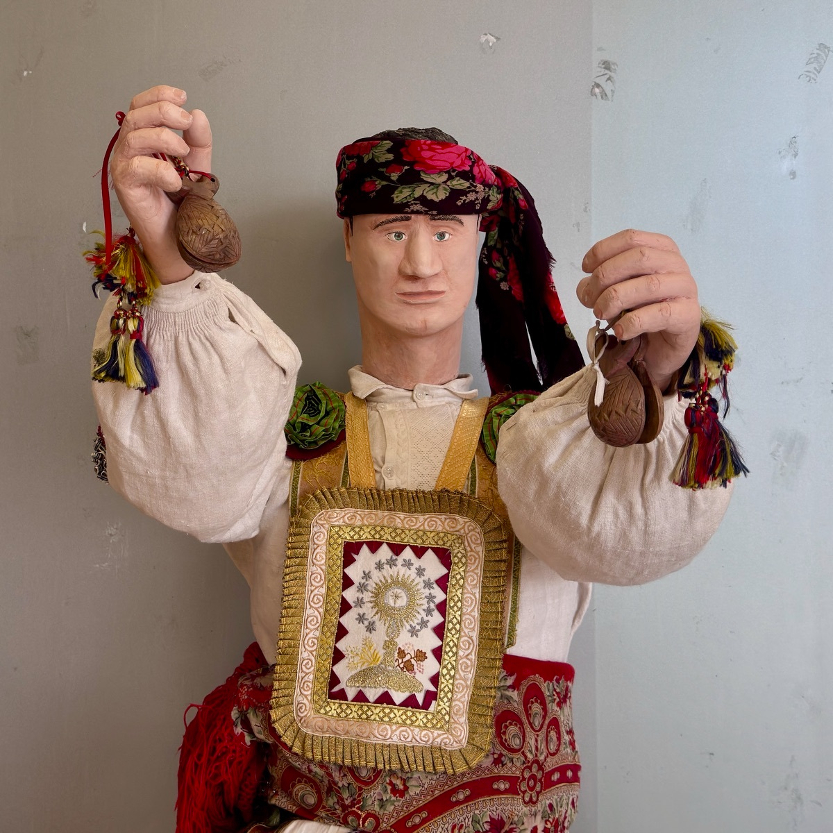 Folk-art figure of a man in traditional Iberian costume - white blouson sleeves, embroidered scapular with a chalice motif, floral red headscarf - holding wooden castanets aloft in each hand with colorful tassels.