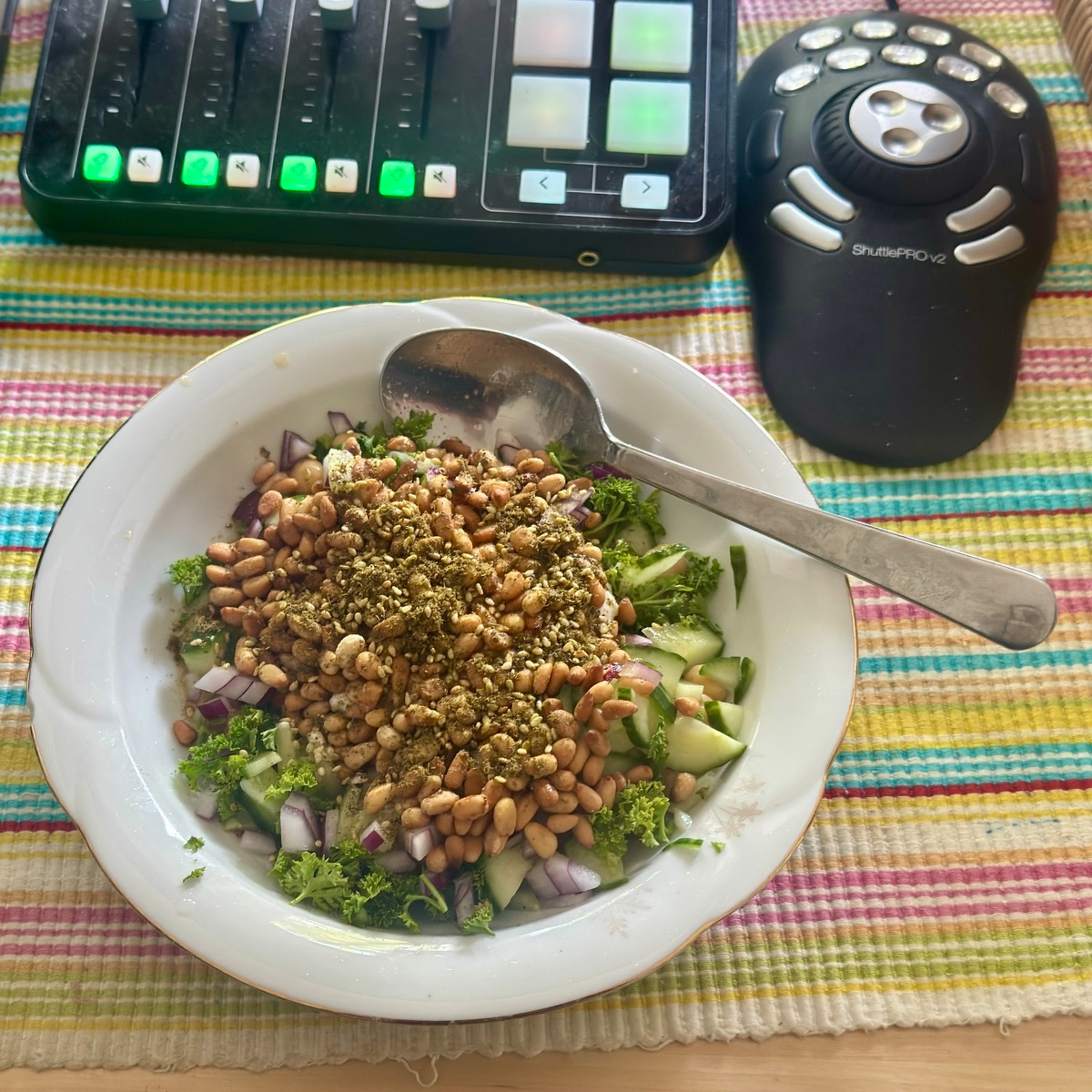 Bowl of green salad topped with a mound of pine nuts and za'atar, on a striped placemat next to a Stream Deck and a ShuttlePRO v2 controller.