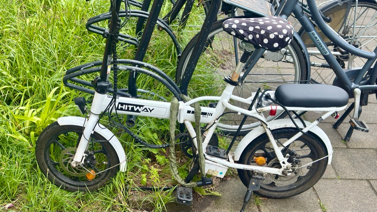 A scuffed white HITWAY folding e-bike chained up beside a black Batavus, parked on a tiled sidewalk against tall grass.