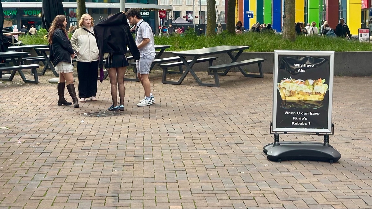 Four people chat on a brick-paved square next to picnic tables, beside a sandwich-board ad asking 'Why have Abs when U can have Kurio's Kebabs?'