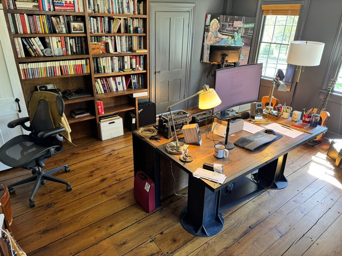 Home office with wood floor, tall bookshelves crowded with books, a wide wooden writing desk holding a monitor, lamp, and papers, and an Aeron-style office chair on the left