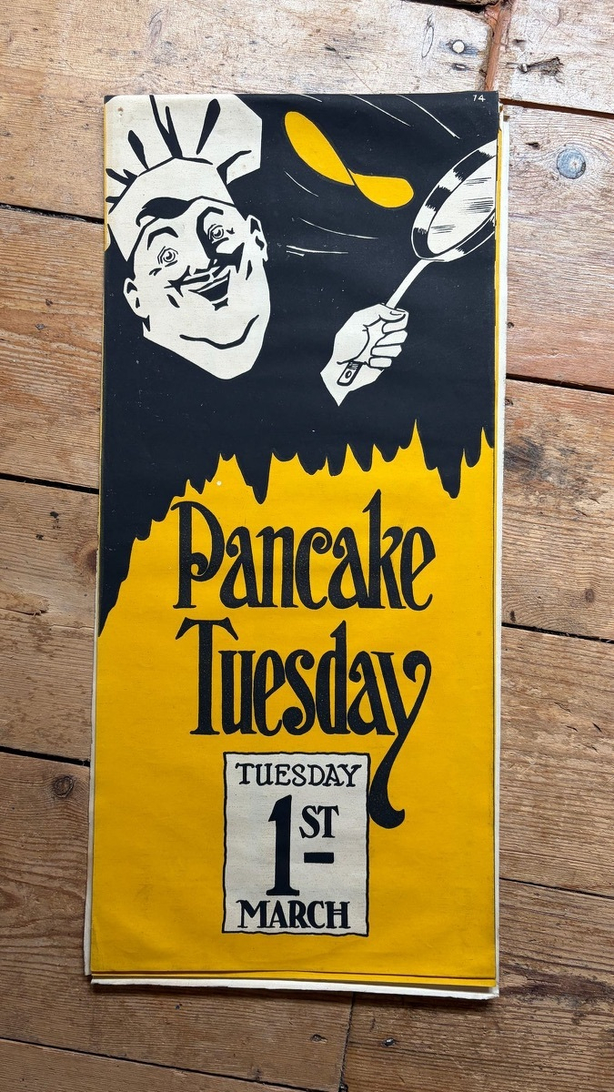 Vintage yellow-and-black 'Pancake Tuesday' poster: a cartoon chef in tall white hat flips a pancake out of a frying pan, with date label 'TUESDAY 1ST MARCH'