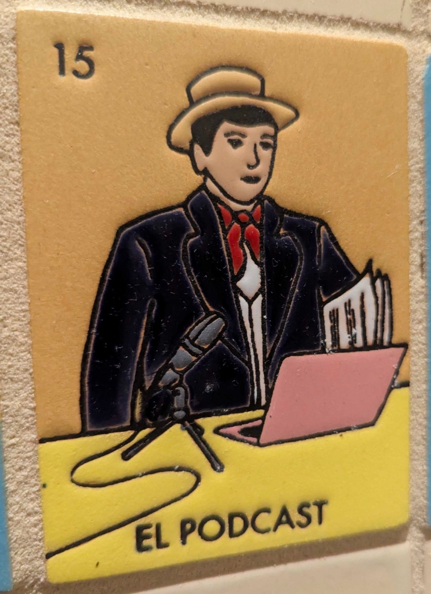 Loteria-style card numbered 15, illustrated with a man in a straw hat, dark jacket and red bow tie speaking into a microphone with a laptop and stack of papers in front of him, labeled 'EL PODCAST'