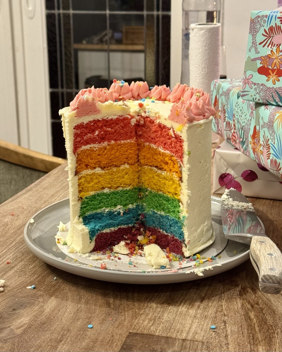 A tall rainbow layer cake with a slice removed, revealing six colorful layers from red to purple, topped with pink rosettes, sitting on a gray plate with birthday gifts in the background.
