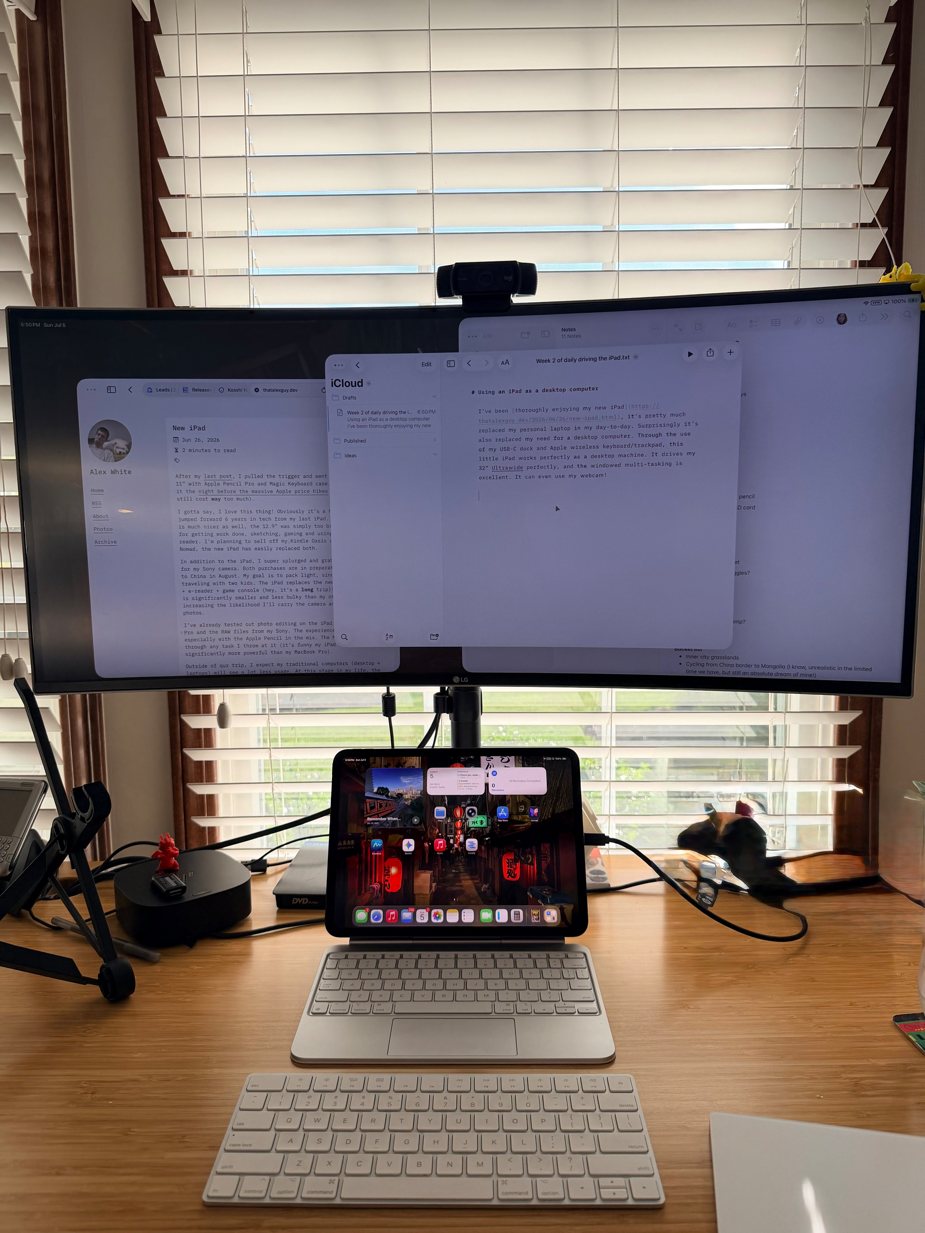 A dual-screen workspace setup with a large monitor displaying text documents, an iPad showing app icons, a keyboard in front, and a cat playing near the window.