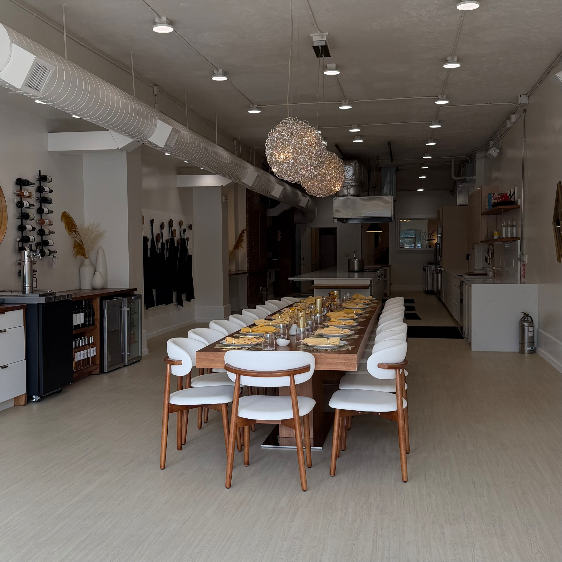 A team-building space ready with a long wooden table set with plates, cutlery, and napkins, surrounded by white cushioned chairs, with two artistic pendant lights hanging above.