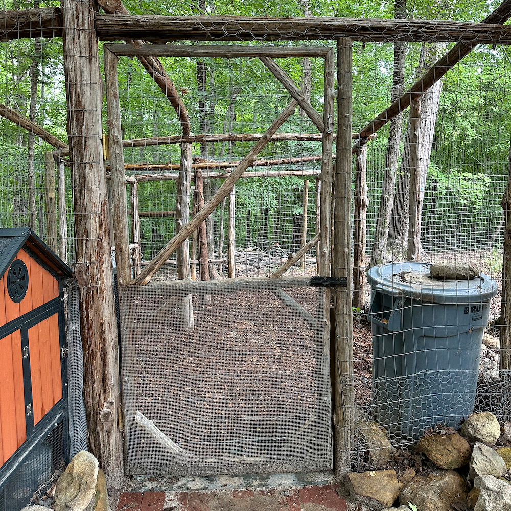 Auto-generated description: A rustic outdoor enclosure with a wooden frame, wire mesh gate, and a trash bin is situated next to a small wooden structure in a wooded area.