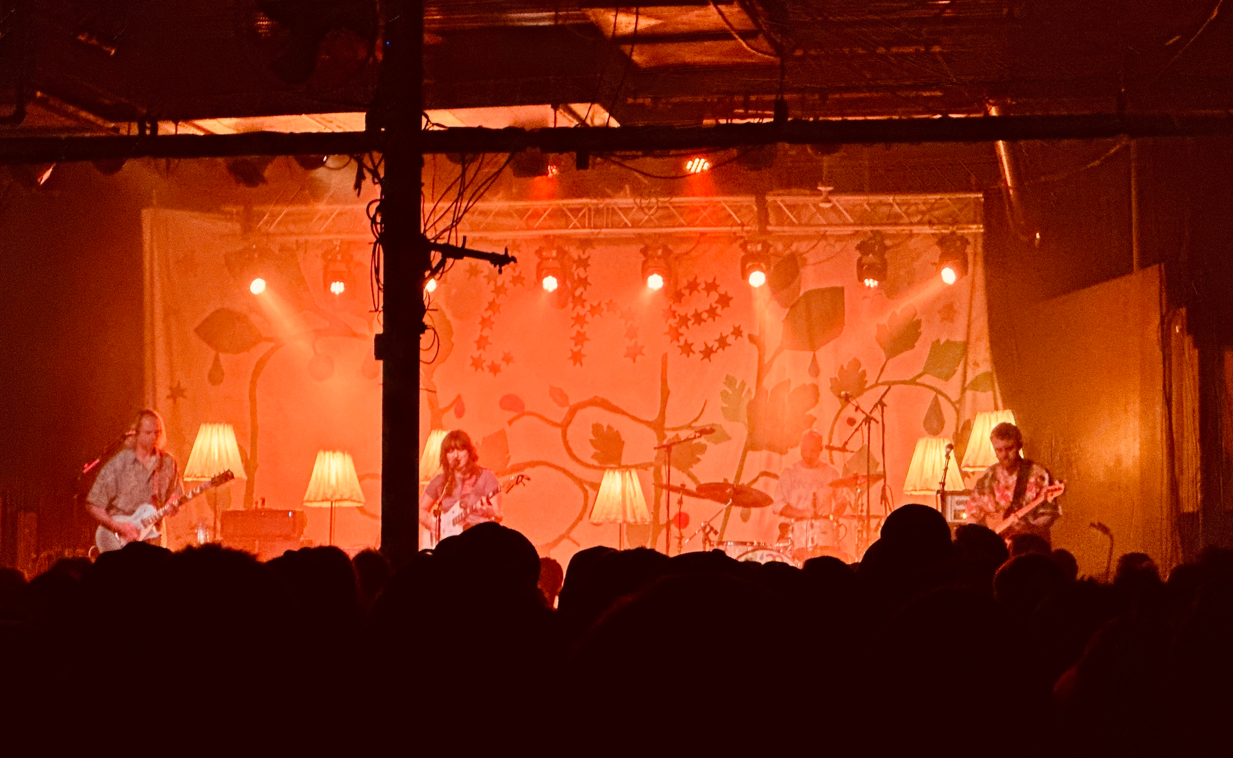 Auto-generated description: A band is performing on a stage with warm orange lighting and decorative lamps, while a crowd watches in the foreground.
