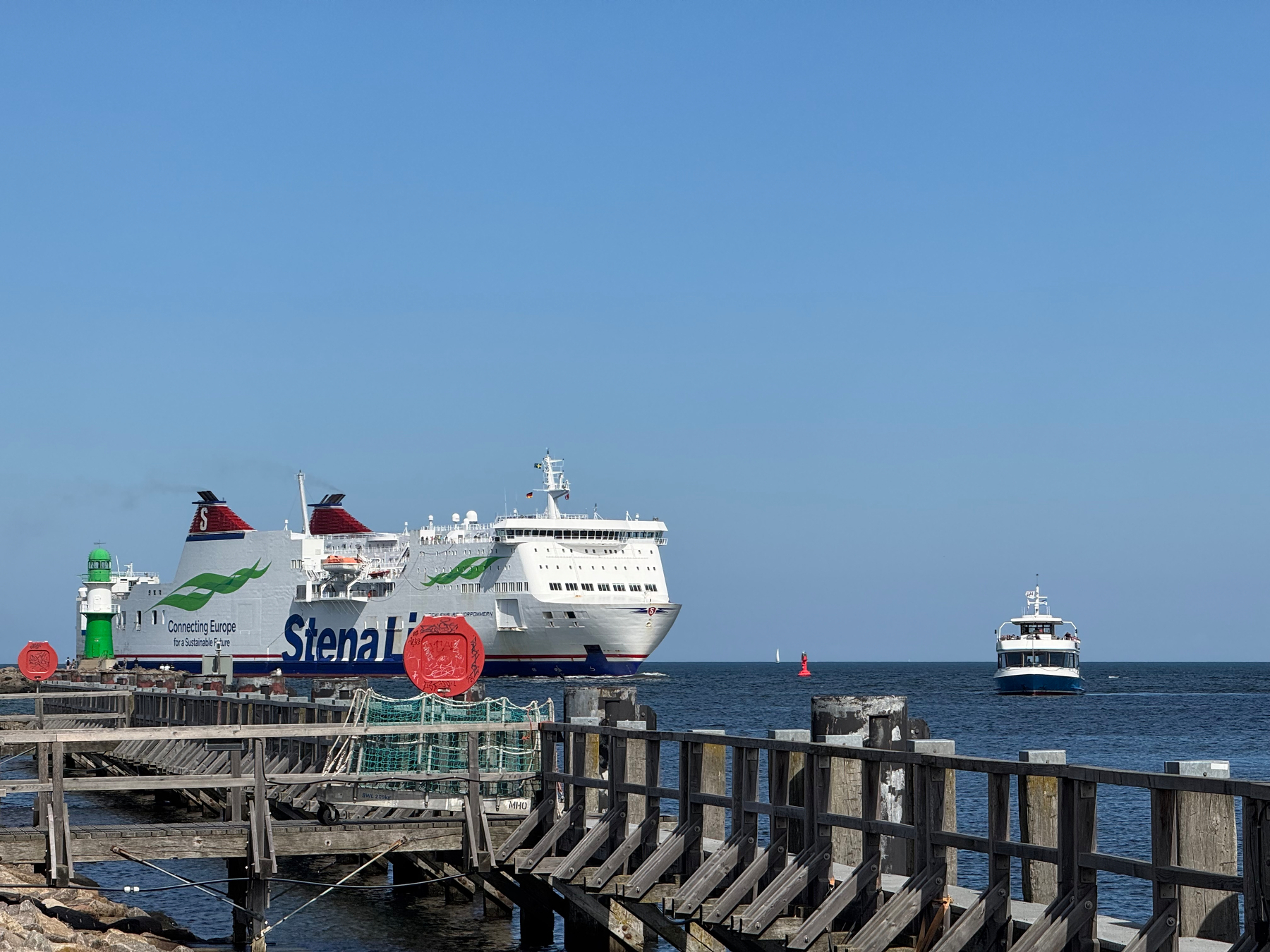 Ein großes Fährschiff der Stendal Lines und ein kleineres Hafenrundfahrtboot sind im Hafen Warnemünde mit einem Holzdock und dem grünen Leuchtturm zu sehen.