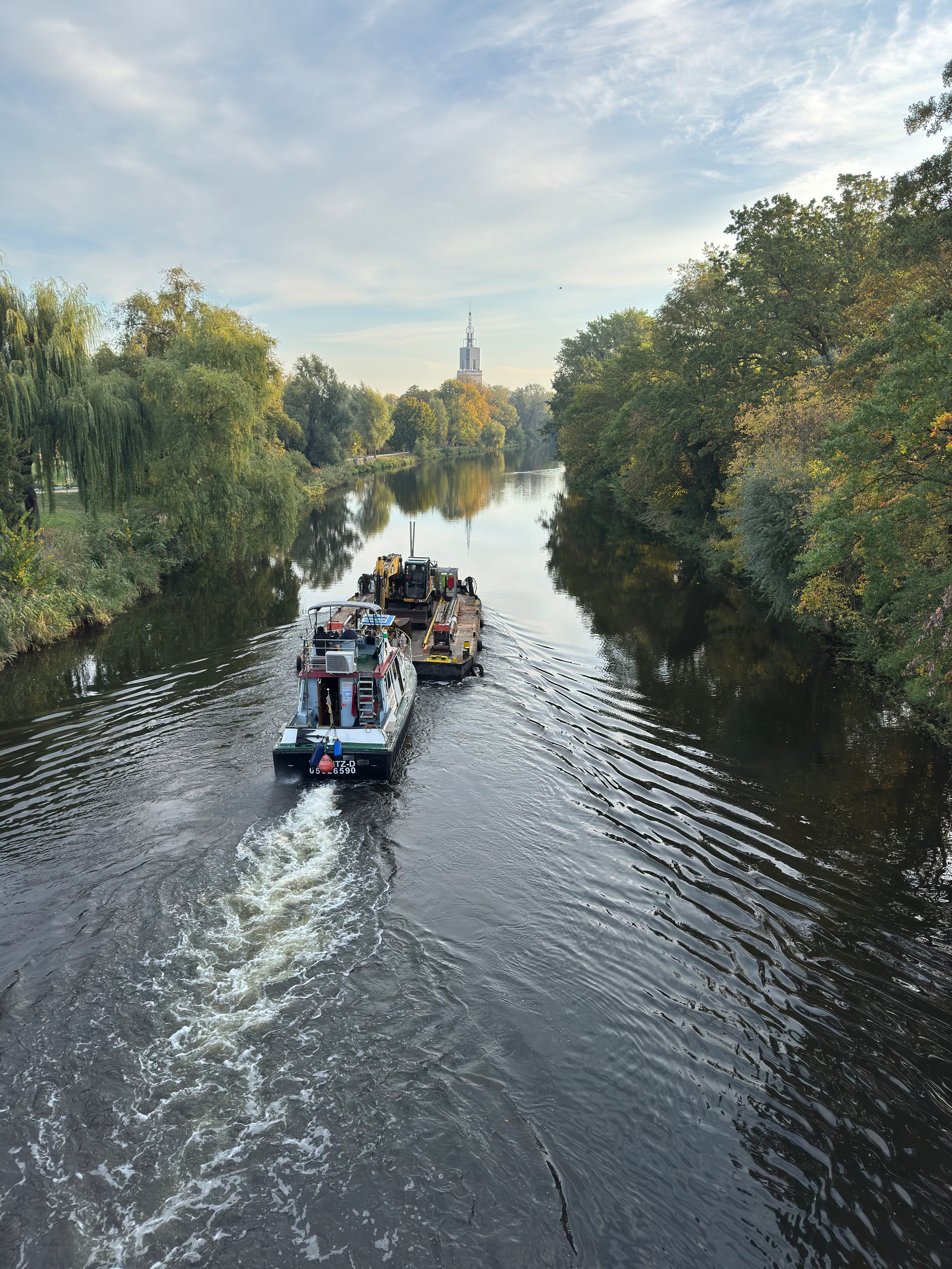 Auf der Havel schiebt ein Boot zieht einen Kahn auf em ein Baufahrzeug transportiert wird. Das Boot ist umgeben von herbstlich bunt gefärbten Bäumen und einem Turm im Hintergrund.