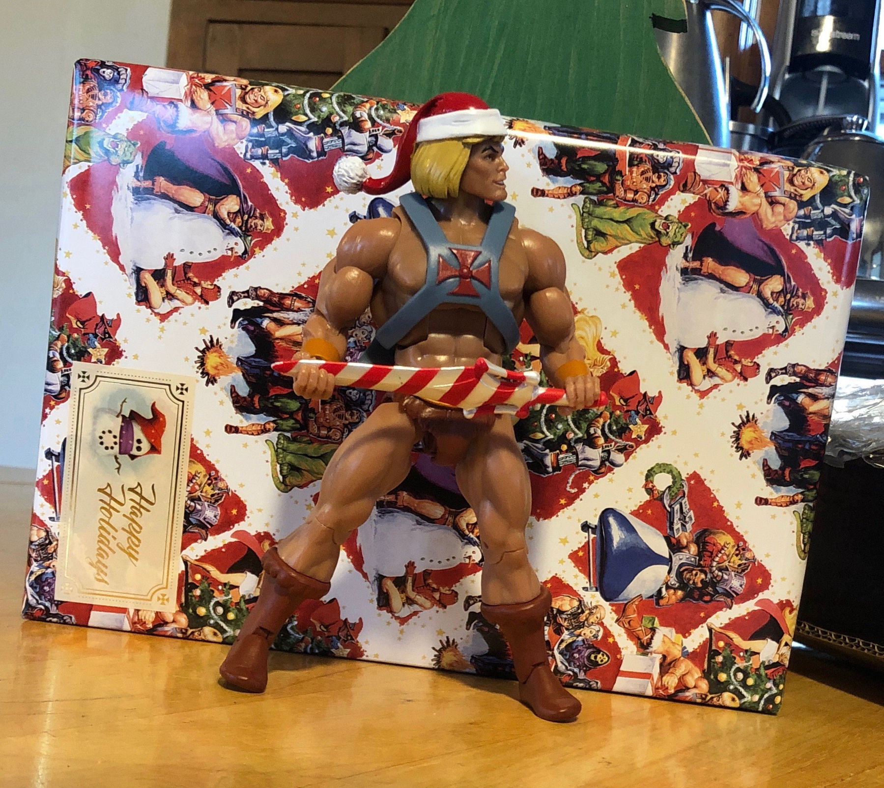 He-Man action figure wearing a Santa hat and holding a candy cane sword in his iconic stance. Behind him is a package wrapped in Masters of the Universe wrapping paper.