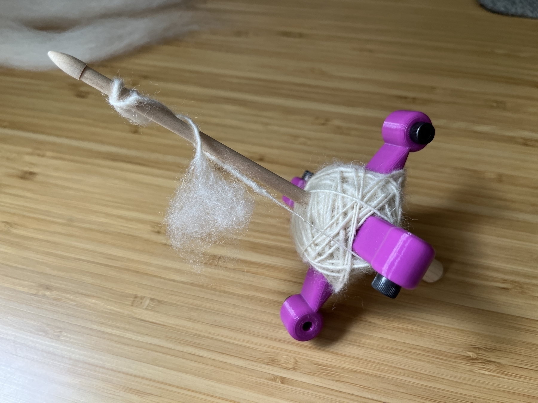 A wooden spindle with 3D printed arms formed into a cross at the bottom of the spindle. A ball of white yarn is partially wound on to the crossed arms. 