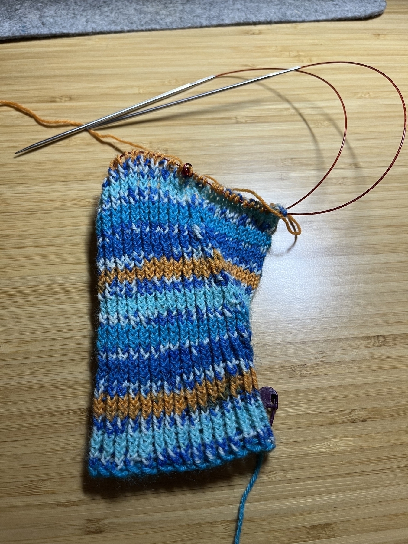 A half-knitted fingerless mitten in self-striping yarn which is the colours of a European Kingfisher. 
