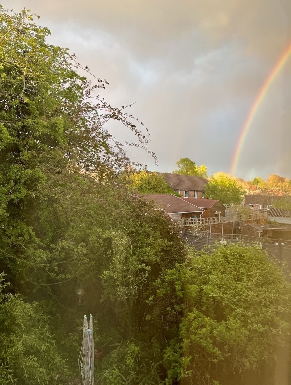 Viewed through a window, a bright rainbow stands out against a dark grey sky to the right of a view of trees and houses.