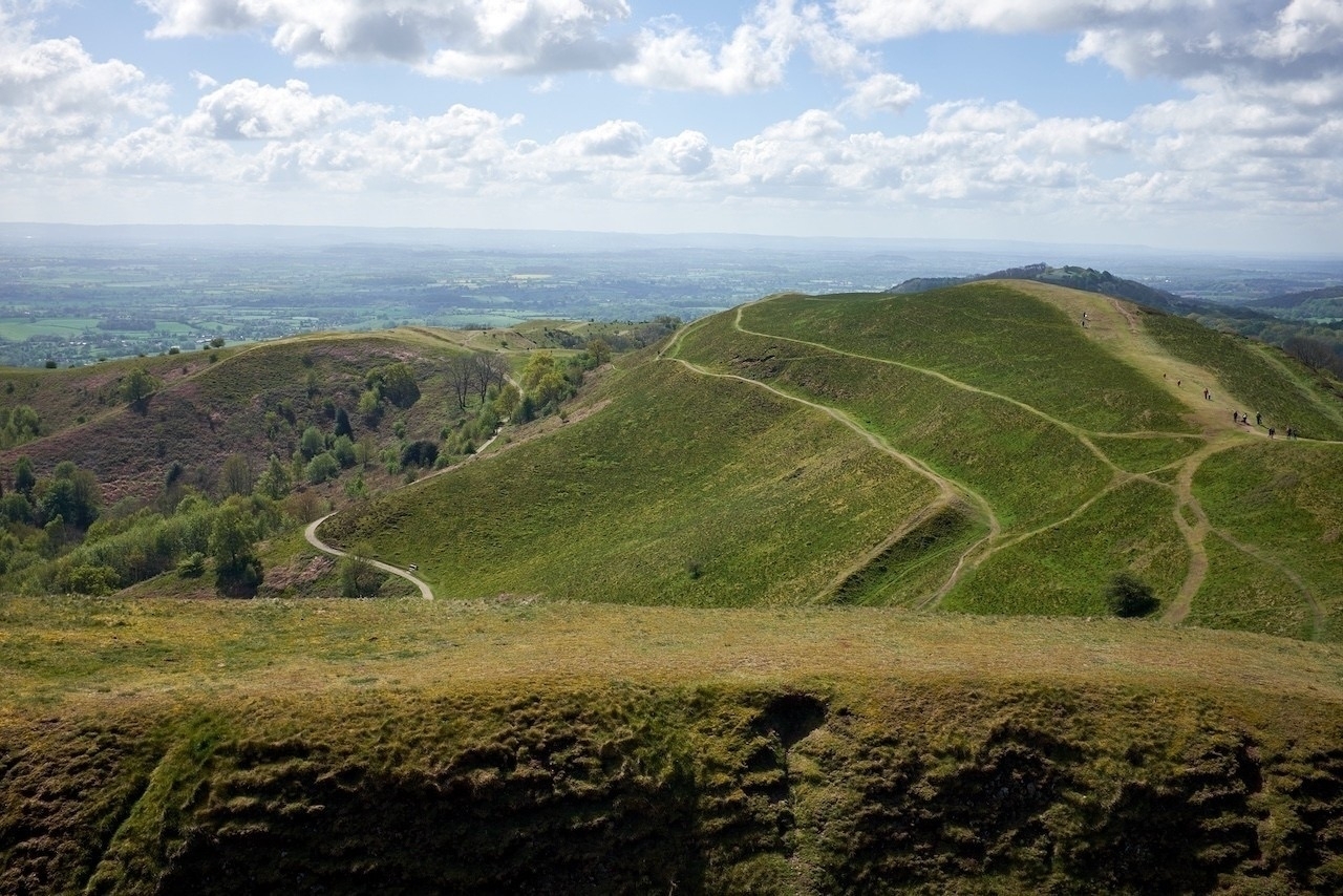Looking across the earthwork ramparts of British Camp Iron Age hill fort in the Malvern Hills. In the middle distance, paths curve around the contours of the rounded hill, and in the distance you can see the flat plains which the Malverns rise dramatically out of.