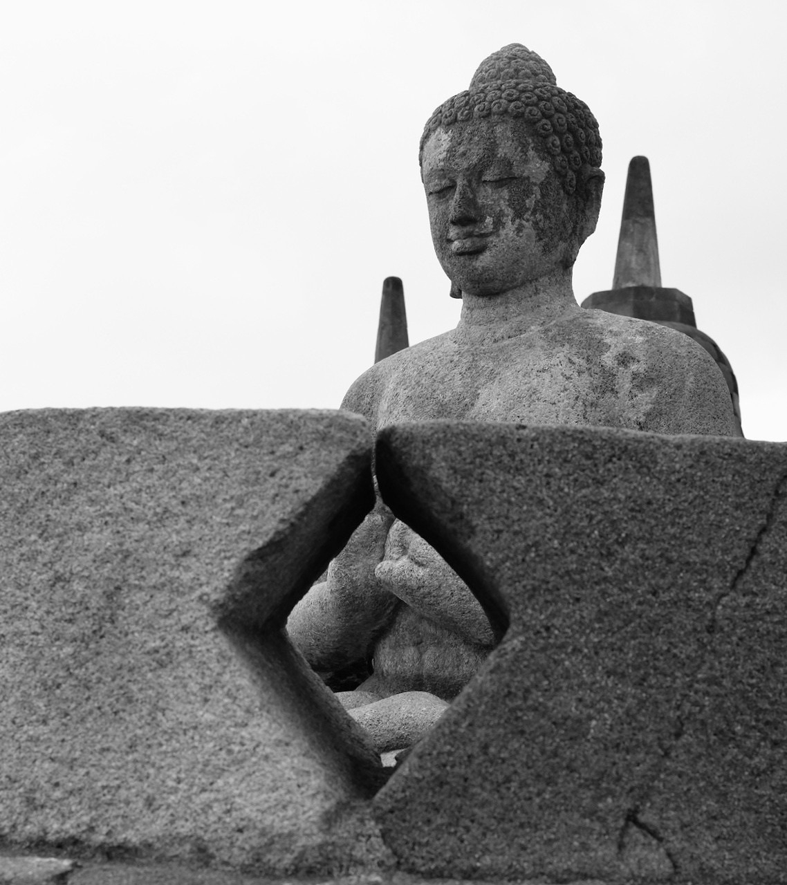 A monochrome photo of a Buddha statue inside a partially open stupa (perforated stone enclosure in an inverted bell shape) at Borobudur Temple, Indonesia. His eyes are closed, and through a perforation in the stupa, you can see the position of his hands in a particular mudra.