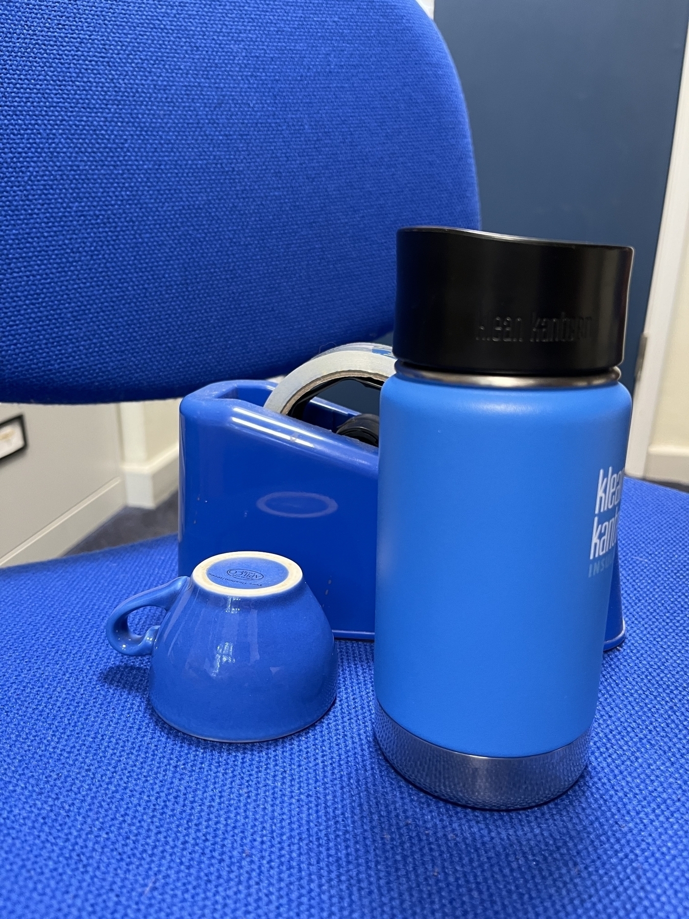 A group of blue objects on a blue office chair, with a blue door in the background: a tape dispenser, an espresso cup and a thermal travel mug.