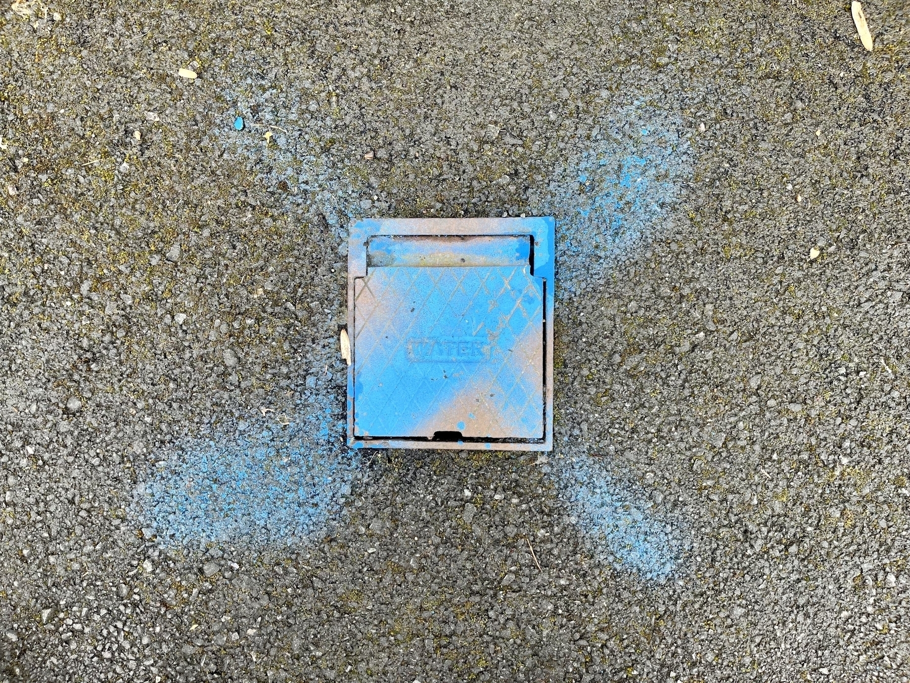 A small metal access hatch for a mains water stopcock in a pavement, with a blue cross spray painted over it. 