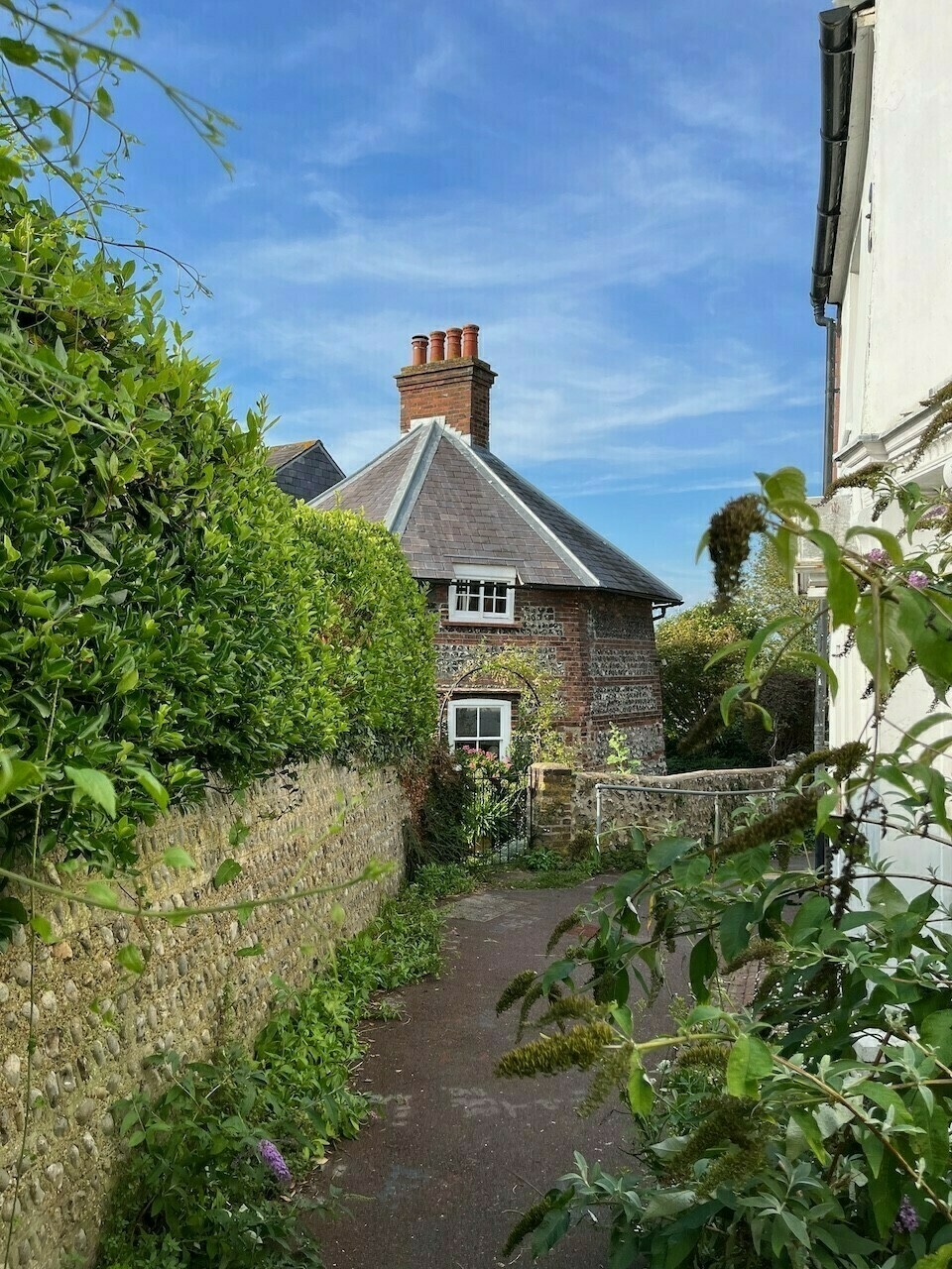 View down a narrow alley (twitten) lined with flowers and a hedge, towards a hexagonal brick and flint cottage, which was once the base of a windmill.
