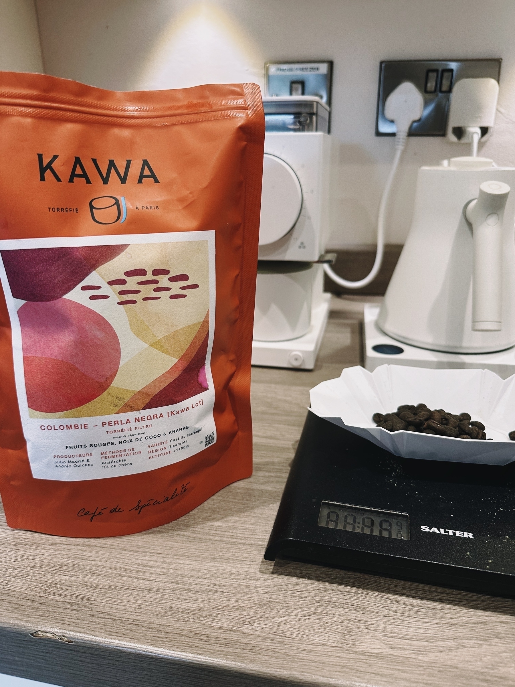 The photo shows a bag of Kawa coffee placed on a kitchen counter. The bag is orange and labeled with "Kawa," indicating it contains Colombian Perla Negra coffee beans. In the background, there is a white coffee machine and an electric kettle. In the foreground, coffee beans are placed on a white paper filter resting on a digital scale, which displays a reading of "0.00."