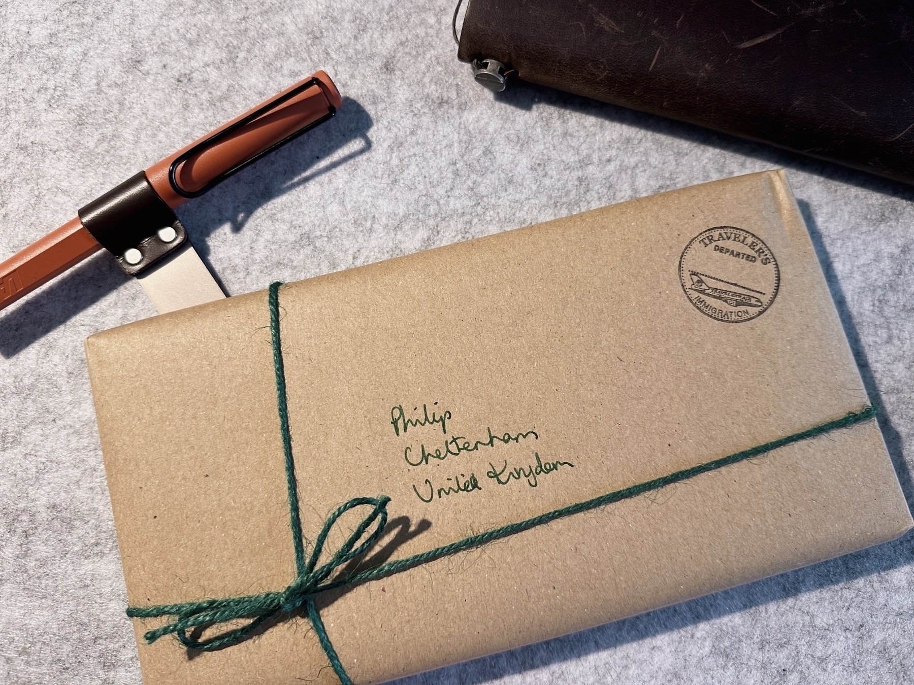 A package wrapped in brown paper with green string sits on a felt desk mat next to a brown travellers notebook and an orange pen in a leather pen loop.
