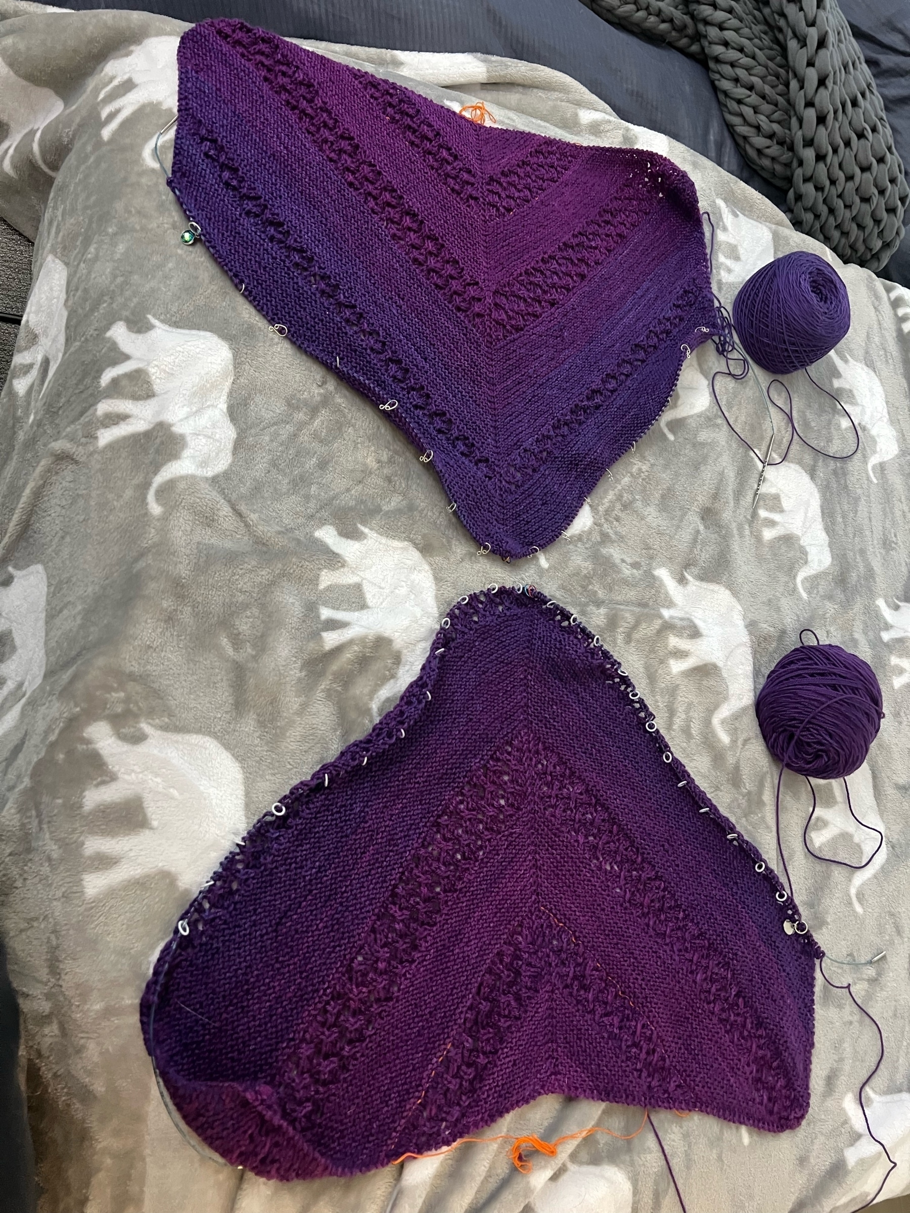 Two largeish trianglular purple knitted things on a bed, points facing one anither.
