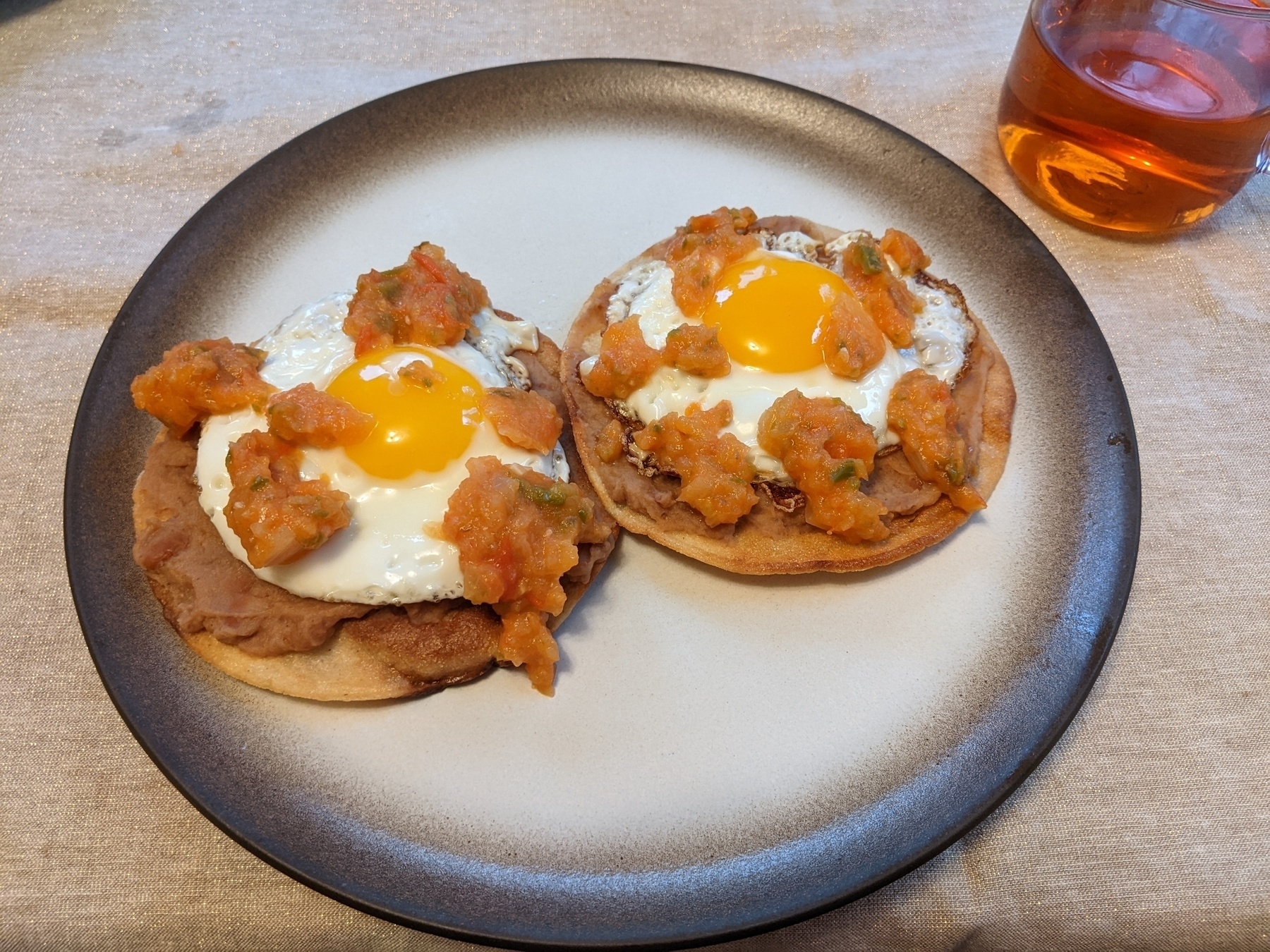 two fried tortillas topped with refried beans, a fried egg, and salsa on a brown and white Heath plate