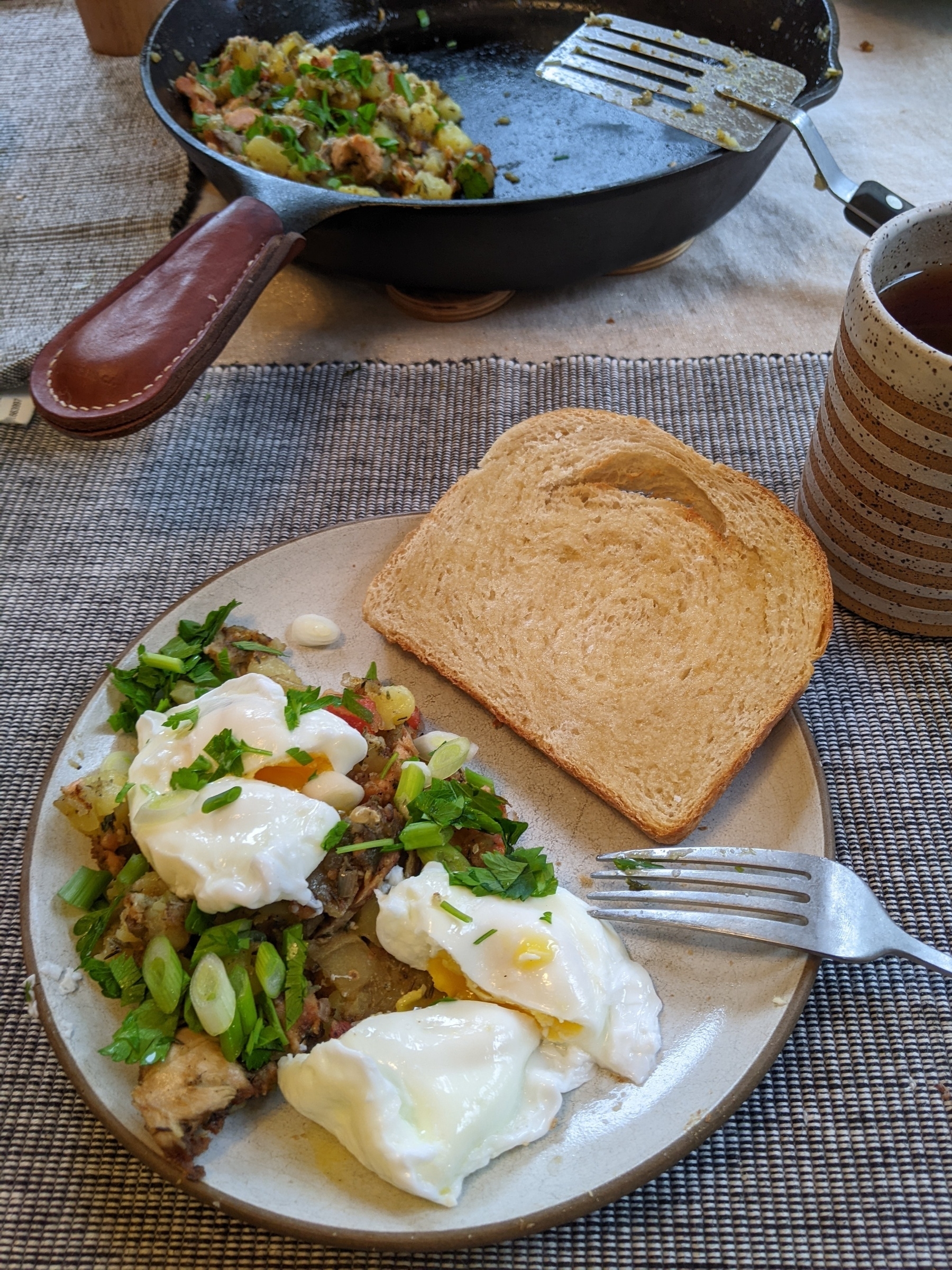 Two poached eggs, split, atop breakfast potatoes with green onions and parsley beside a buttered slice of bread, a cast iron skillet behind 