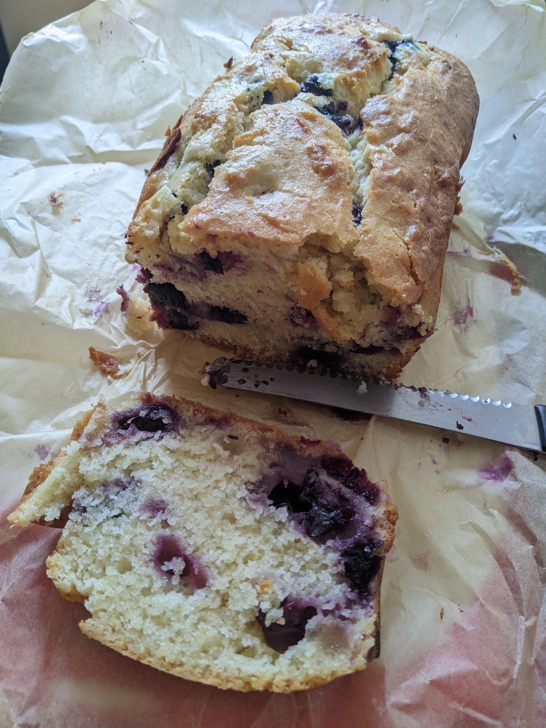 loaf of pale yellow bread spotted with purplish blueberries on parchment paper, a cut slice laying in front 