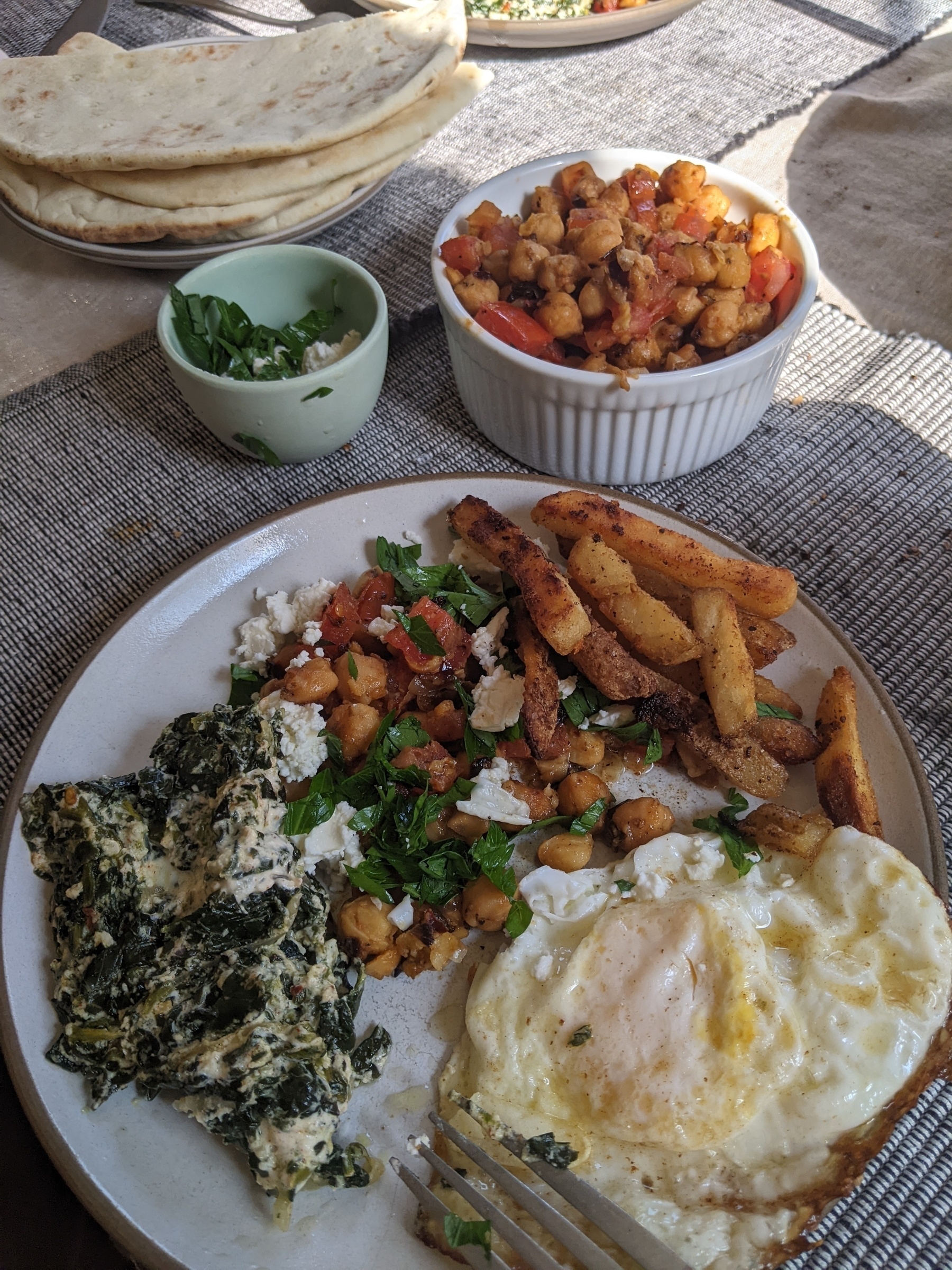 Plate with fried egg, chickpeas topped with feta and parsley, spinach and cream, and refried French fries beside a plate of pita bread 