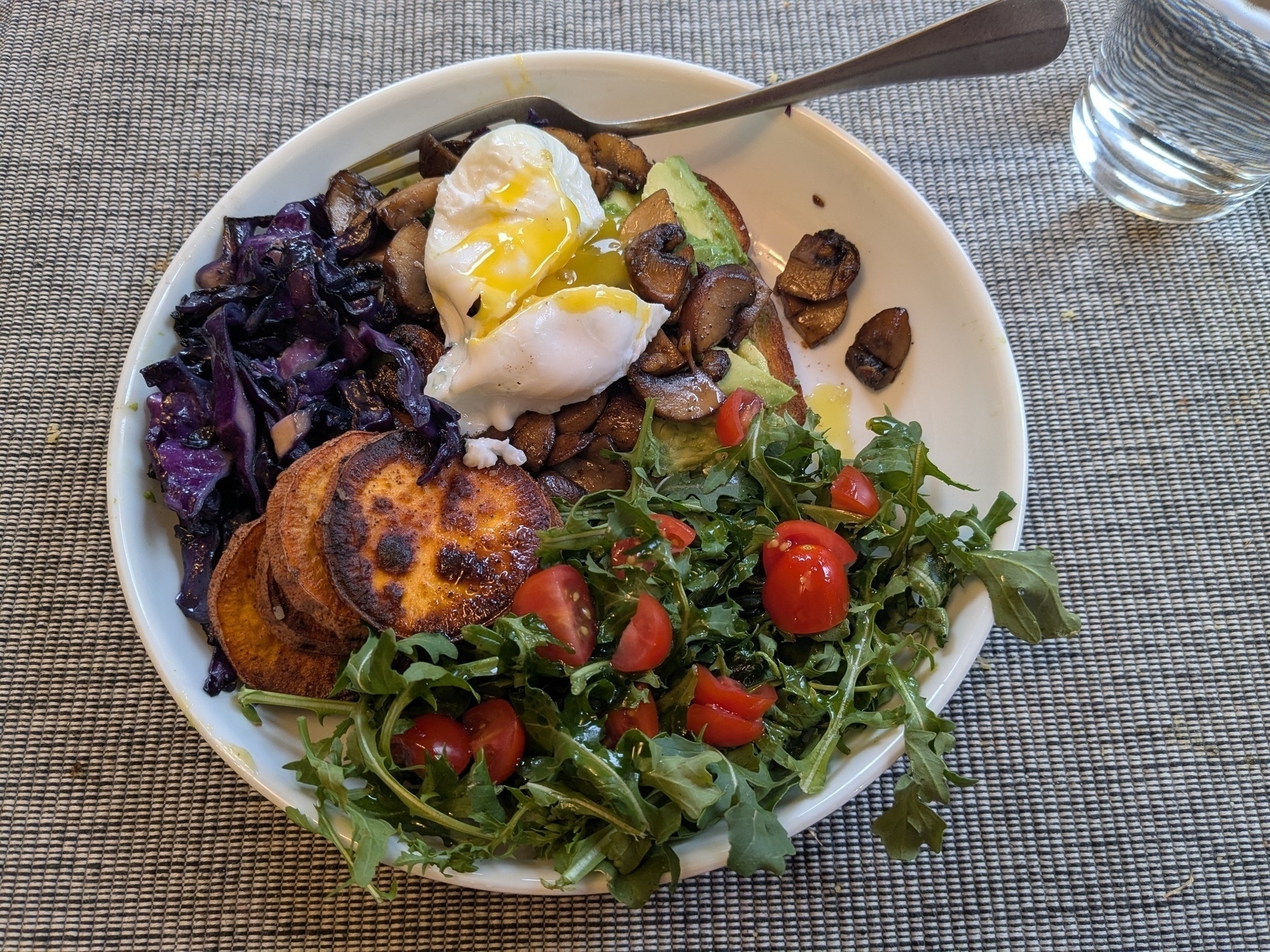 platebowl stuffed with avocado toast topped with a poached egg, a pile of red cabbage, and arugula salad dotted with tomatoes