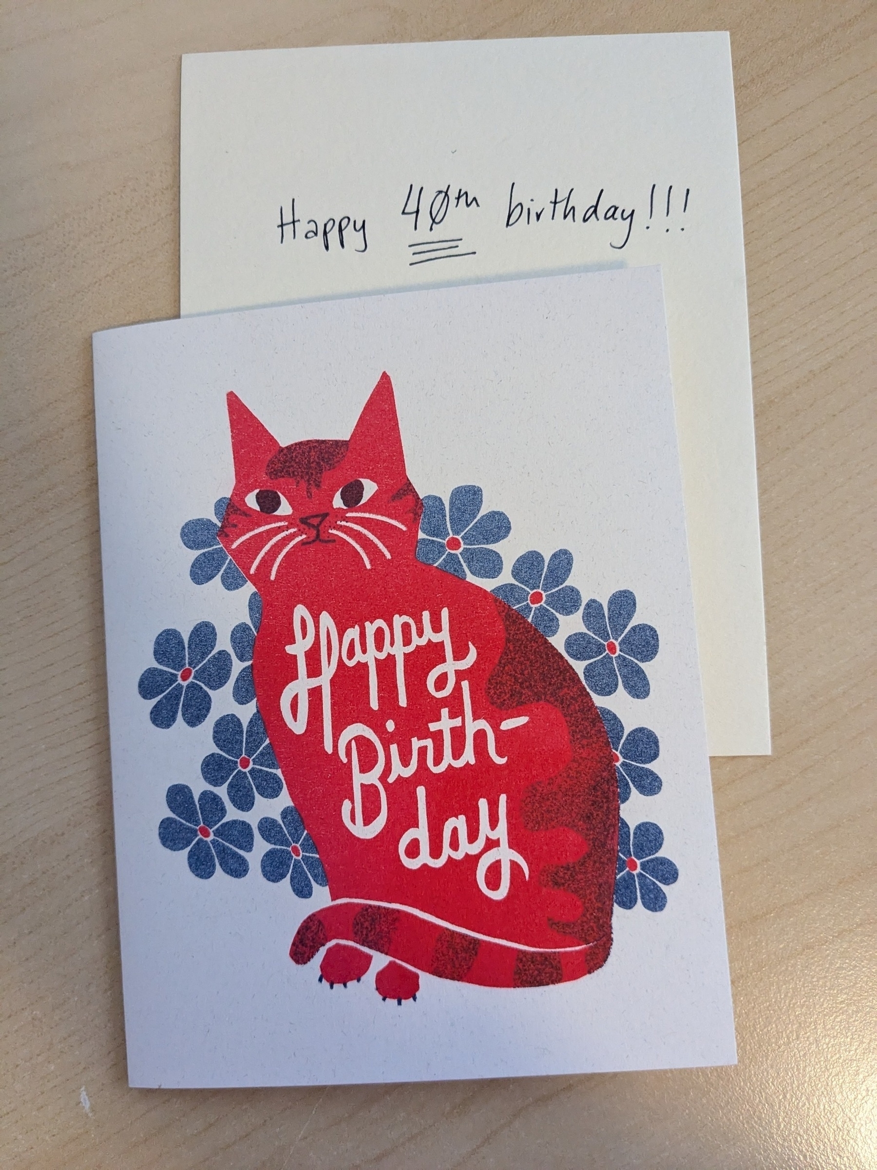 risograph birthday card of red cat in blue flowers saying happy birthday, with a handwritten note on a piece of paper sticking out of the card