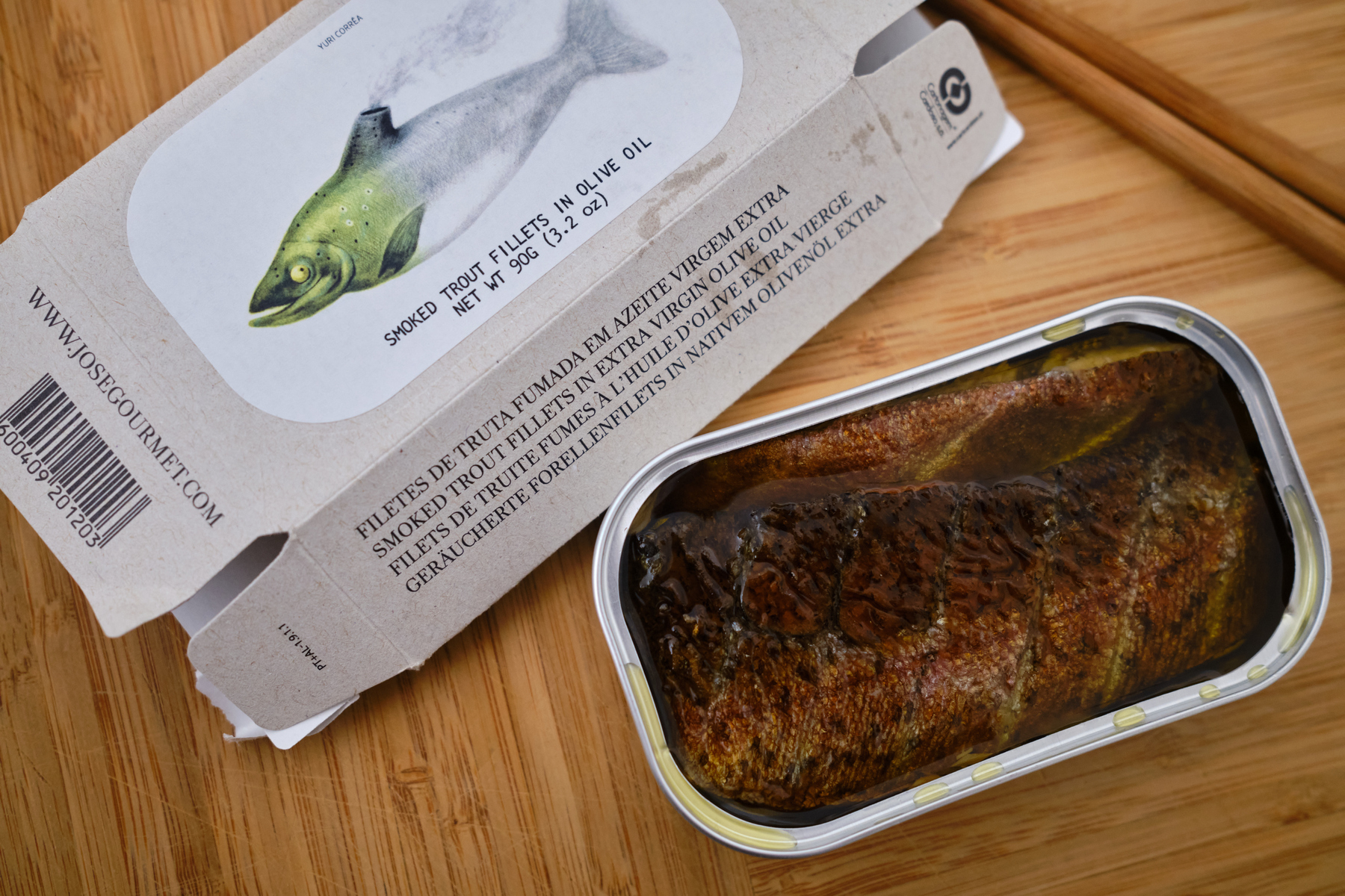 an open rectangular tin of Jose Gourmet's smoked trout in olive oil, positioned next to the art on the package. 