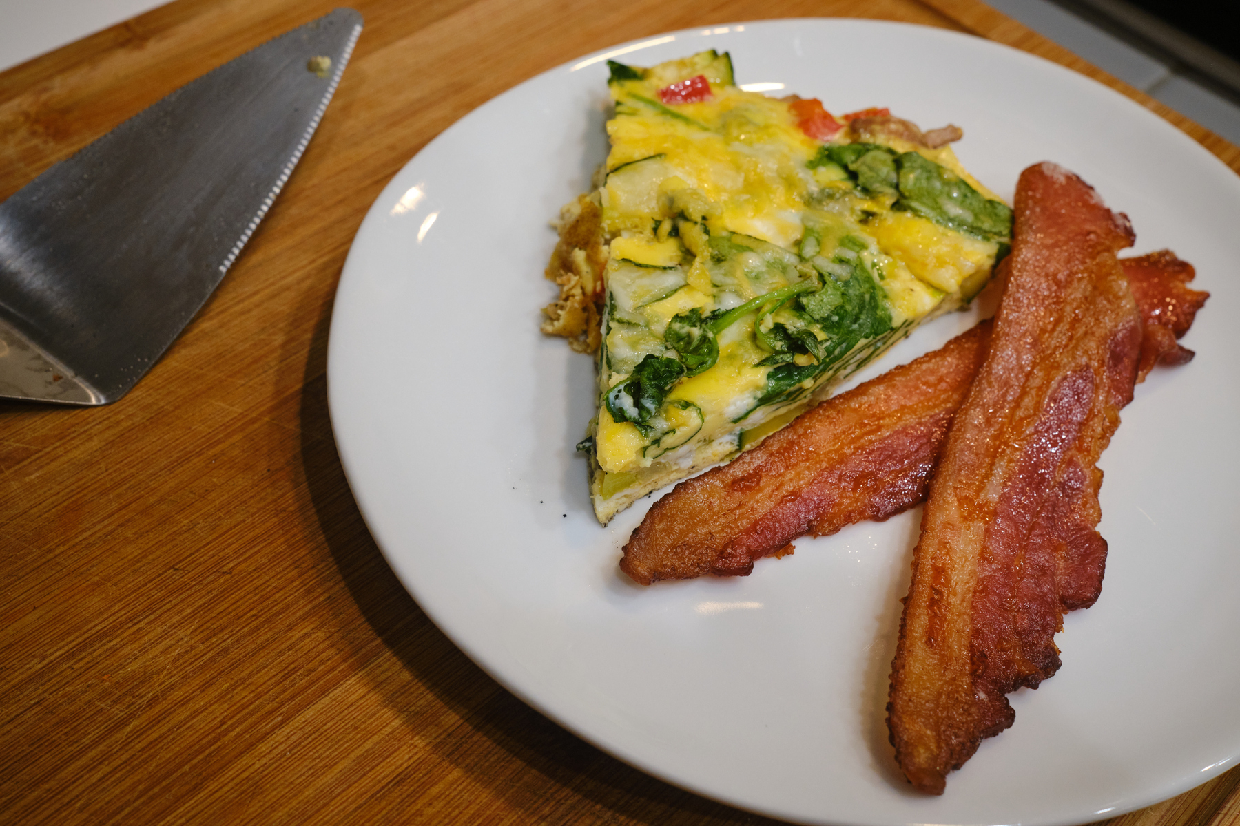 A slice of a fritata on a white plate with two strips of bacon 