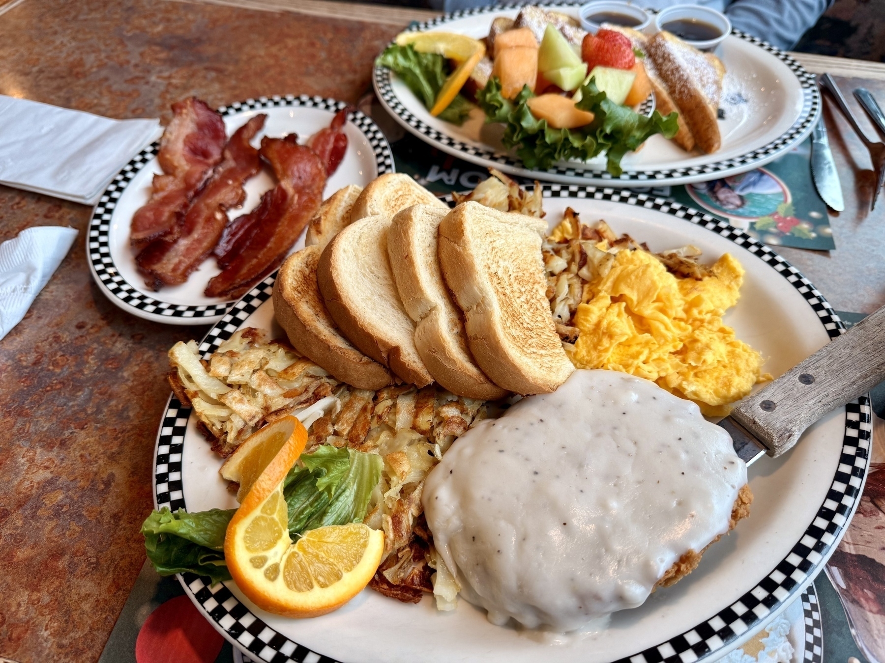 A plate of scrambled eggs and chicken fried steak with hashbrowns and toast. There’s a side of bacon between us, and a plate of French toast for my wife.