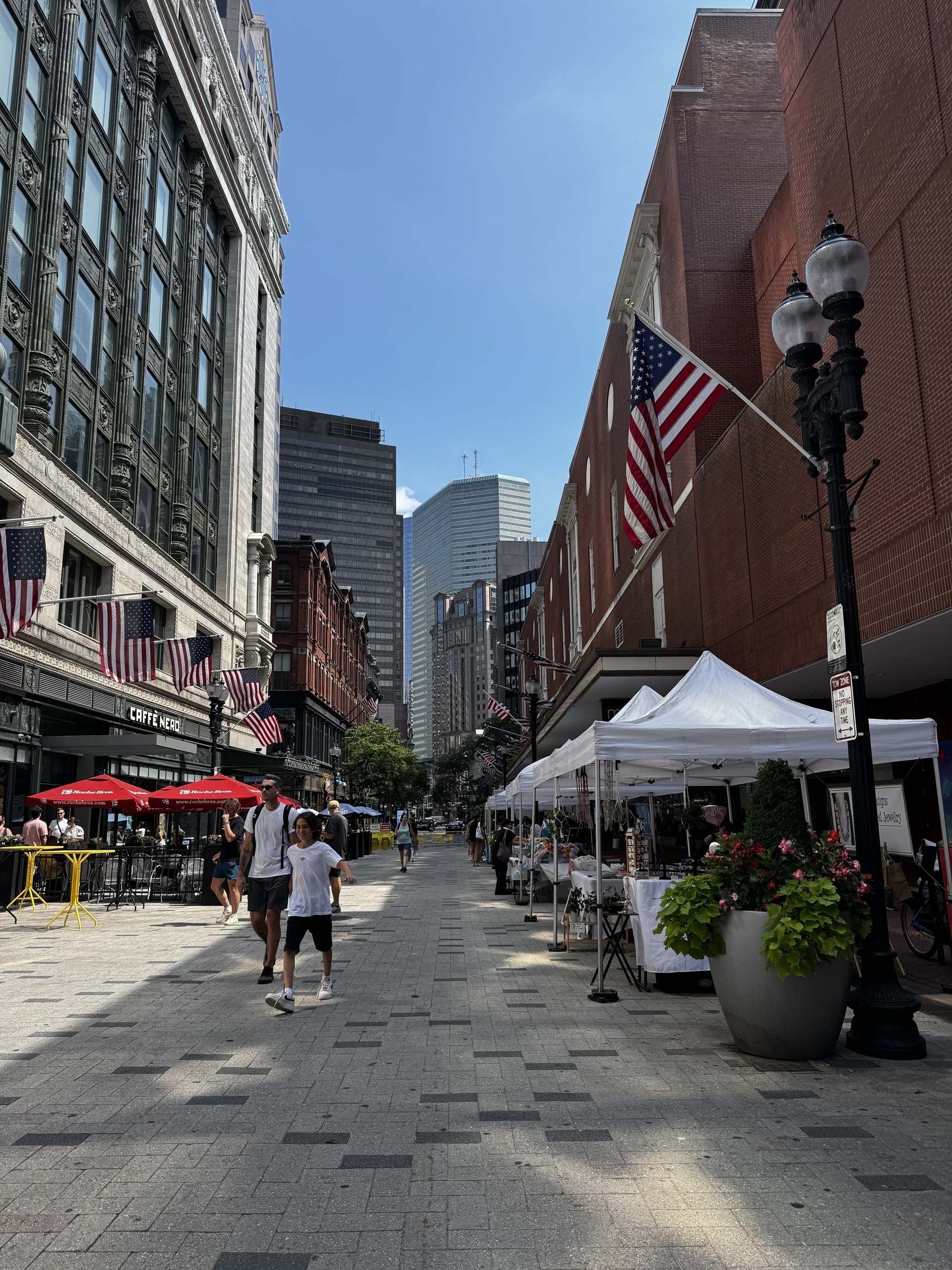 A street level view of a pedestrian way between two large city buildings. In the background, high rises of Boston are visible. An American flag hangs from the right. 