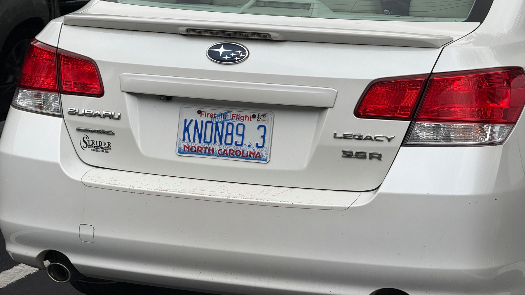 A white Subaru Legacy 3.6R with a North Carolina license plate reading KNON89.3 is parked.