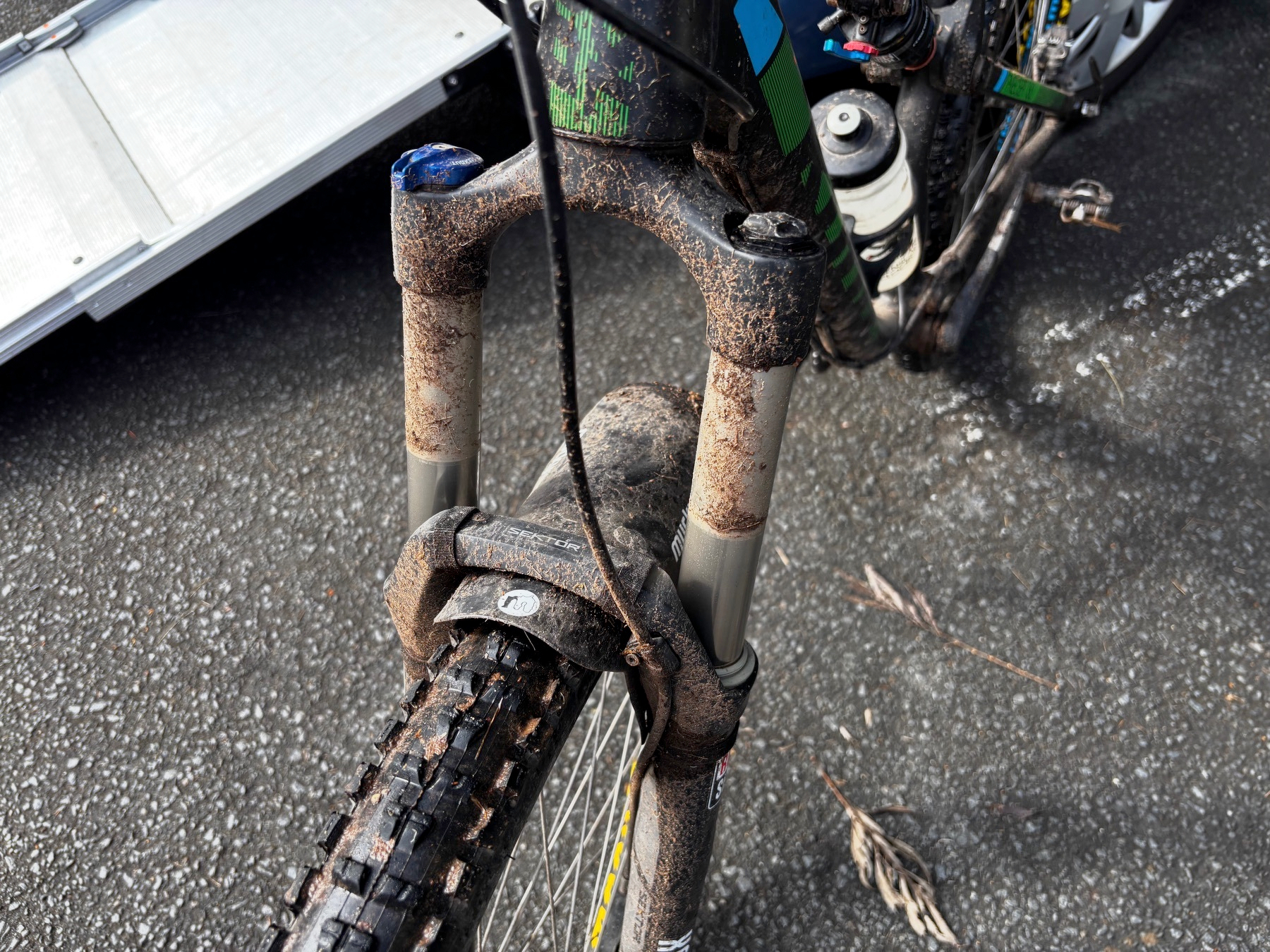 A muddy mountain bike tire and front suspension fork are shown resting on an asphalt surface.