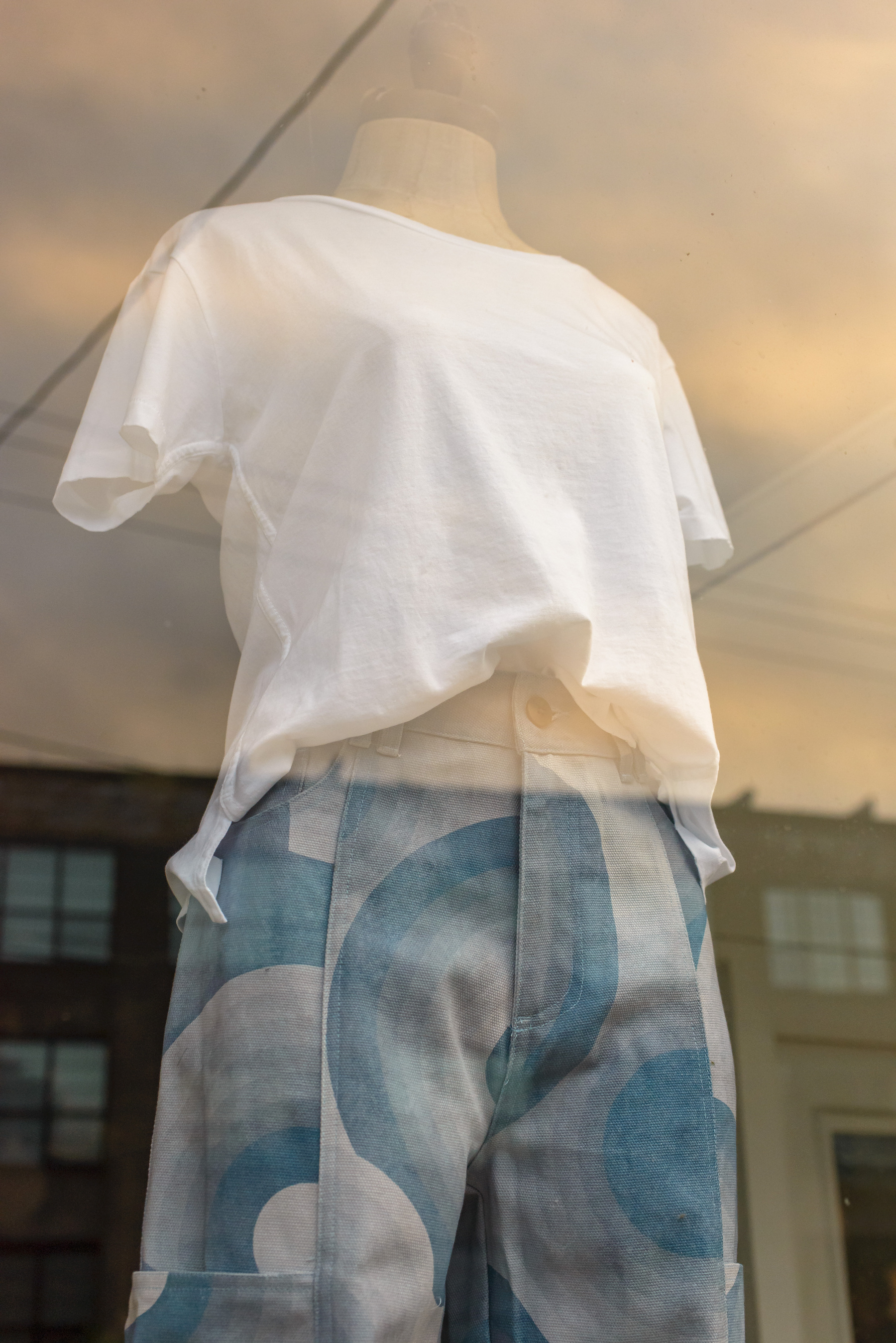 Blouse and pants on a female mannequin in a shop window. Blouse is white cotton knit. Pants are a rainbow circles print with shades of dark to light blue making up the rainbow swirls.