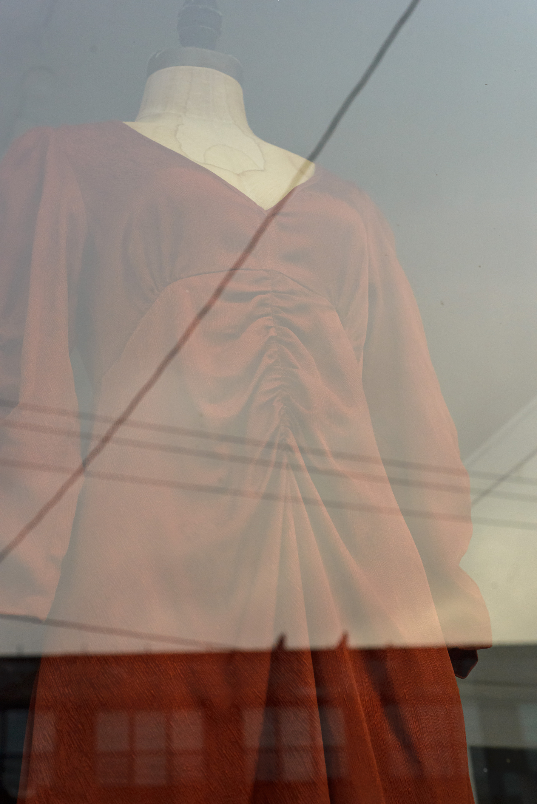Mannequin with rust colored sundress in a shop window with sky, wires and buildings reflected in the window.