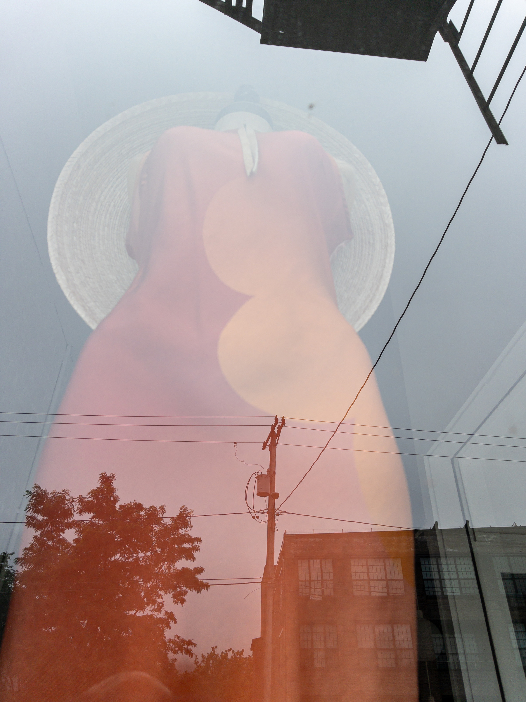 orange and yellow dress and broad brimmed hat on a mannequin in a shop window with sky and buildings reflected on the window.
