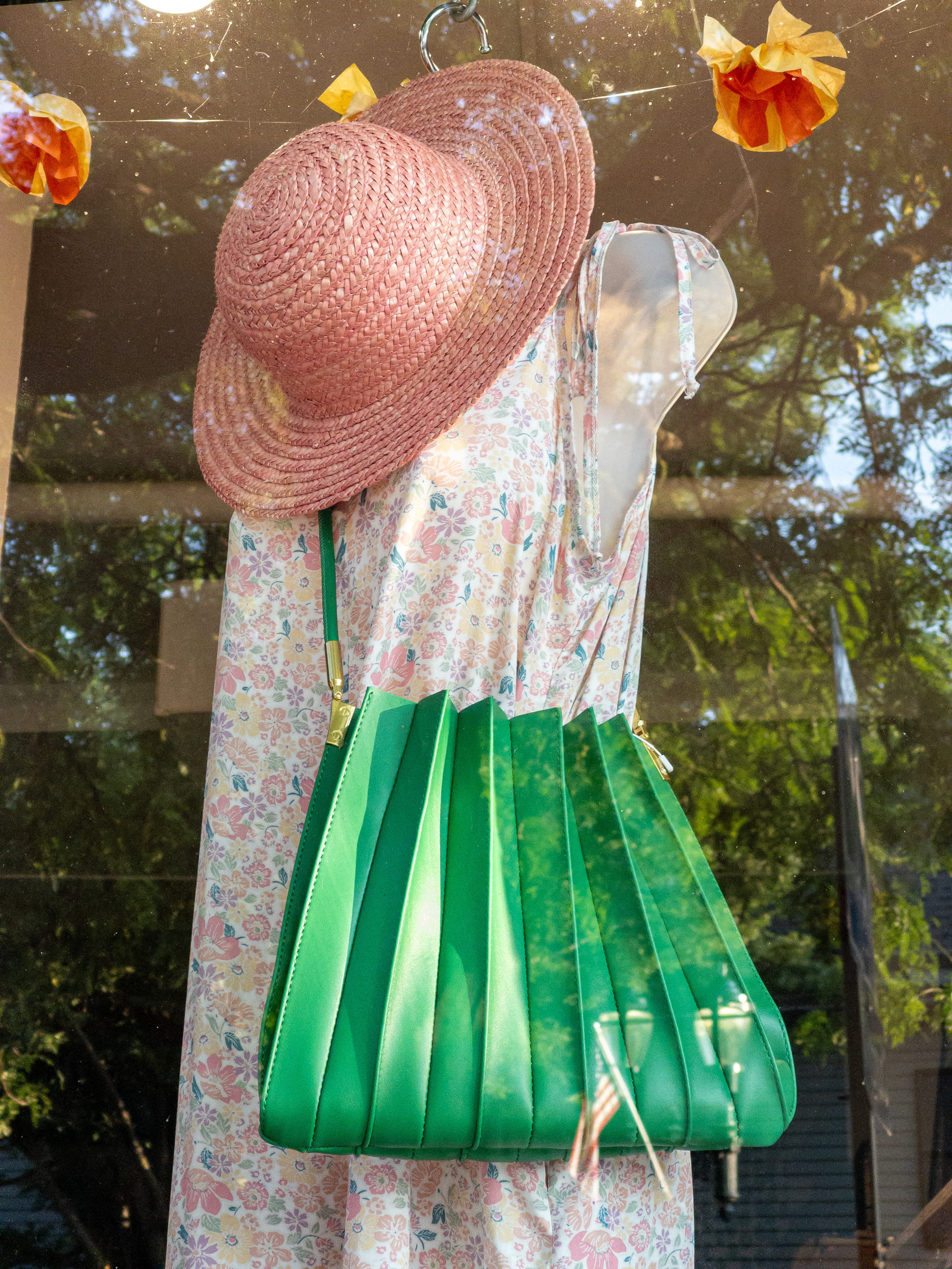 Pink hat and pleated green tote bag/purse draped on mannequin wearing a pink floral sundress in a shop window.