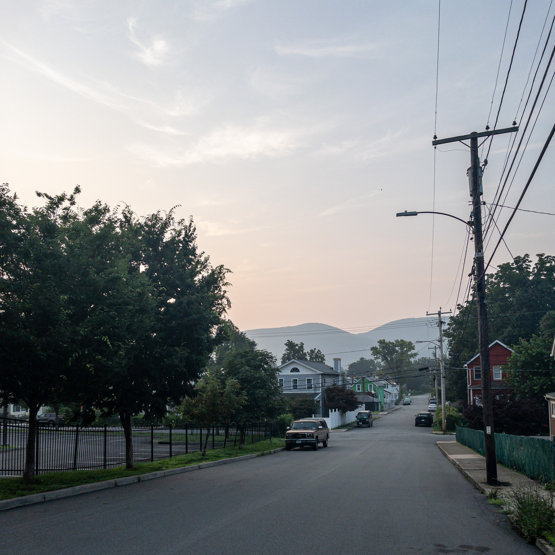 View down a street in the early morning with trees on the left and utility pole and wires on the right side and the hazy outline of rounded mountains in the distance.