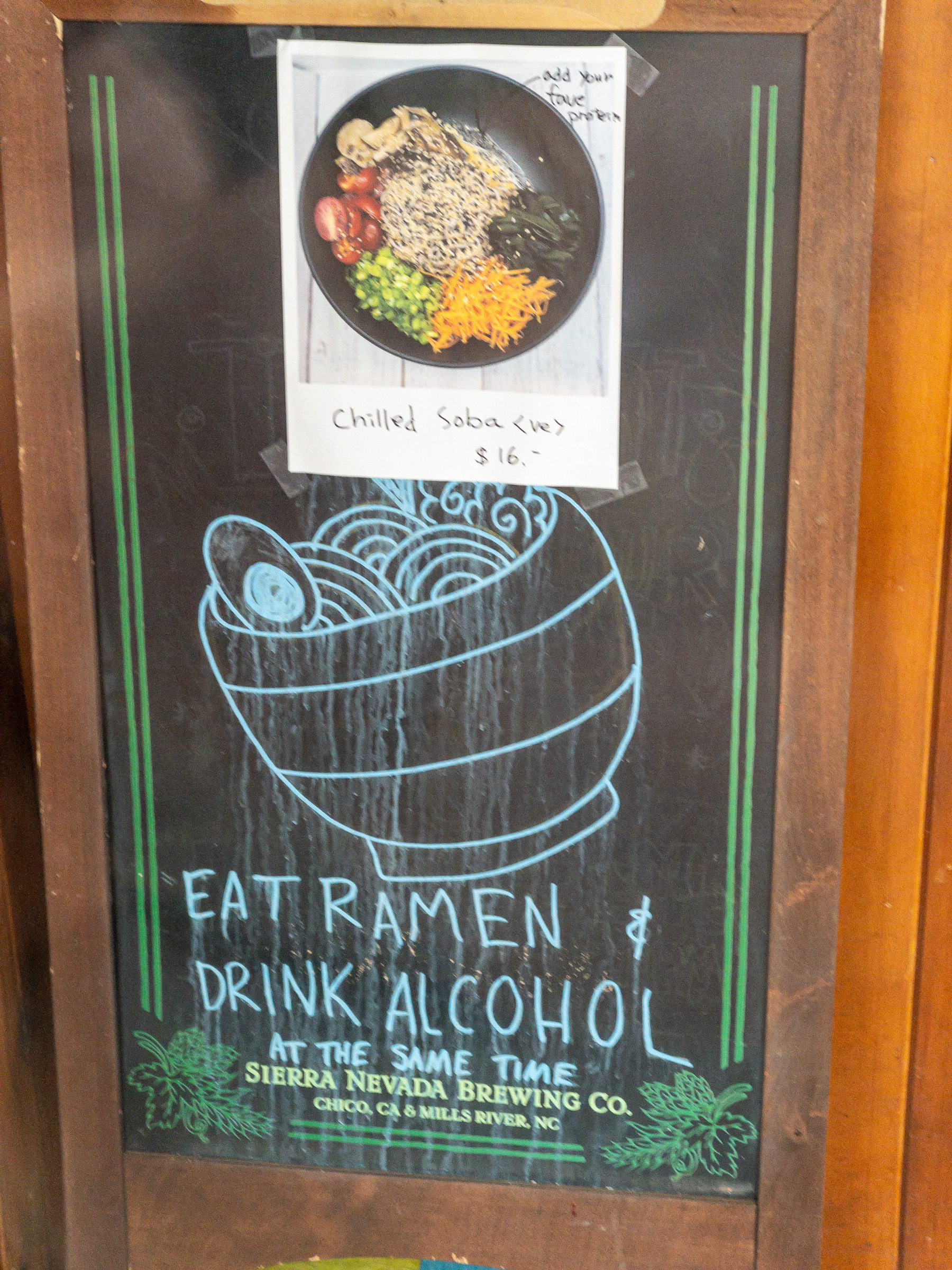 Chalk board sandwich sign with a picture of a bowl of ramen taped to the top portion, a drawing of a bowl of ramen below the picture and the words “EAT RAMEN &amp; DRINK ALCOHOL AT THE SAME TIME” written in capital letters below that.