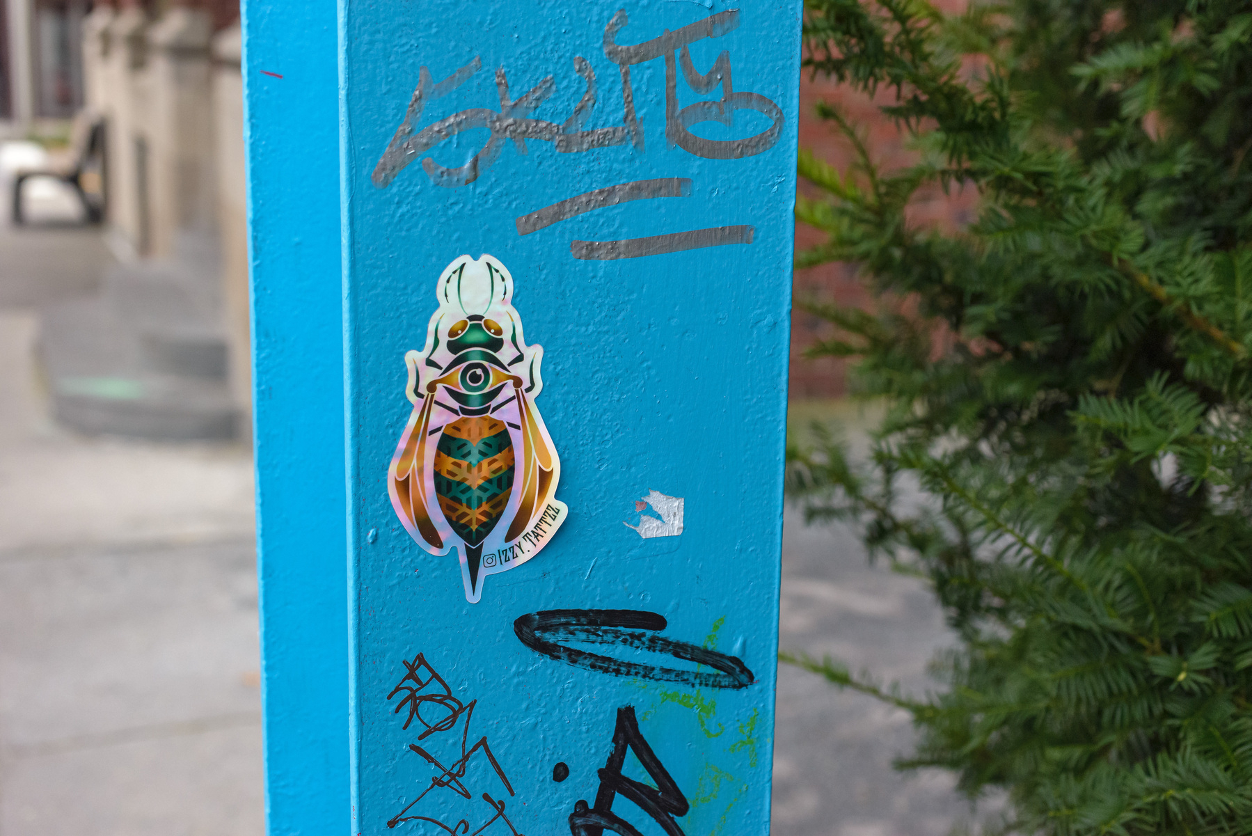 Bug sticker on a steel column painted blue with graffiti. Sticker says IZZY.TATTZZ.
